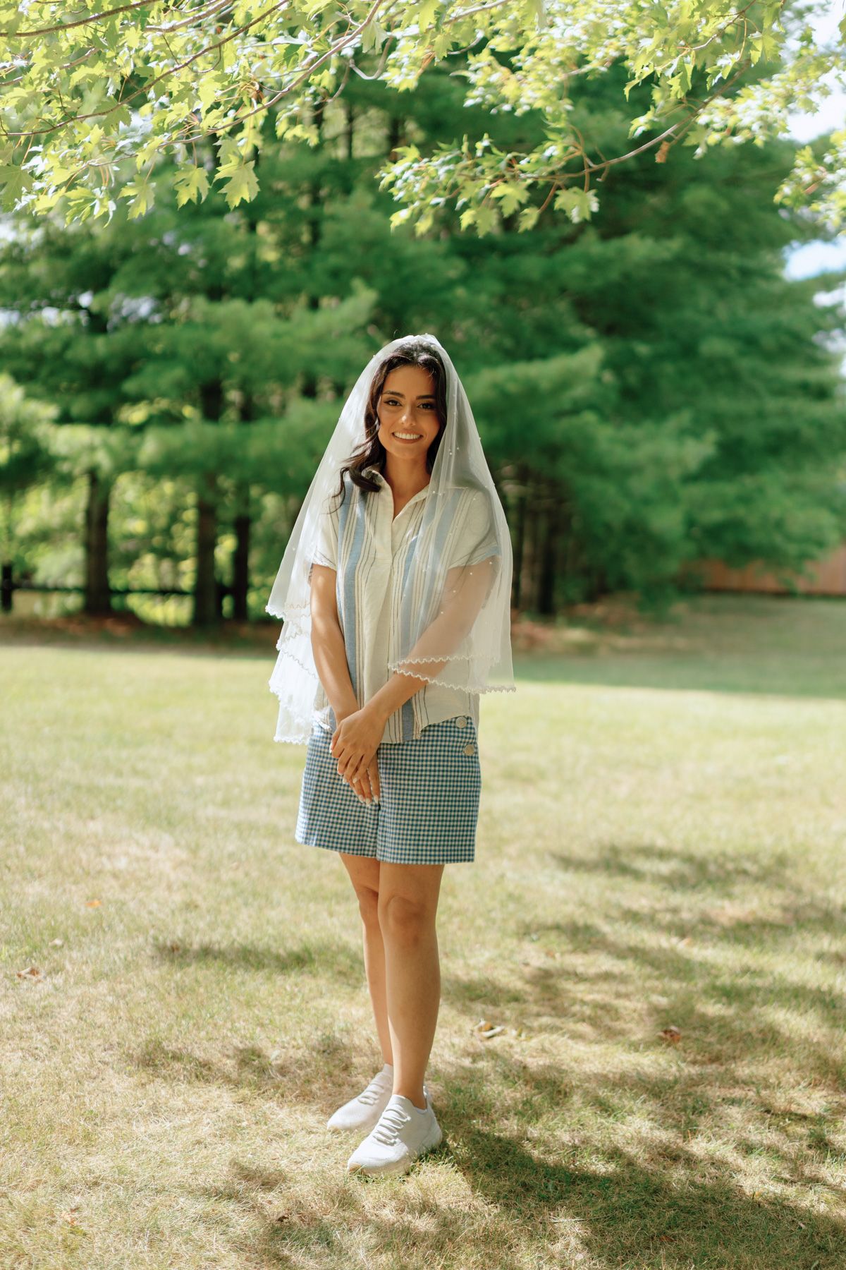 Bride to be in sheer white top and plaid skirt smiling under sunlit trees at Kurtz Orchards in Niagara-on-the-Lake.
