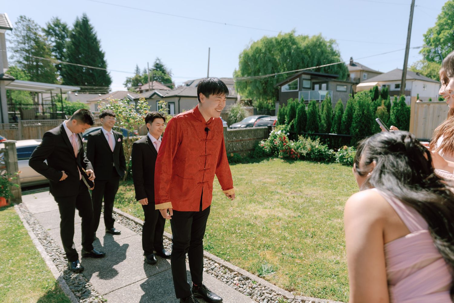 Groom in red traditional Chinese jacket greets guests at an outdoor backyard wedding celebration.