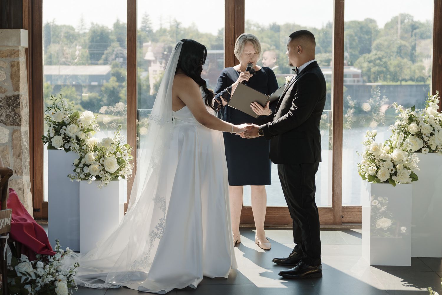 Bride and groom exchanging vows with officiant in elegant venue with white floral arrangements.