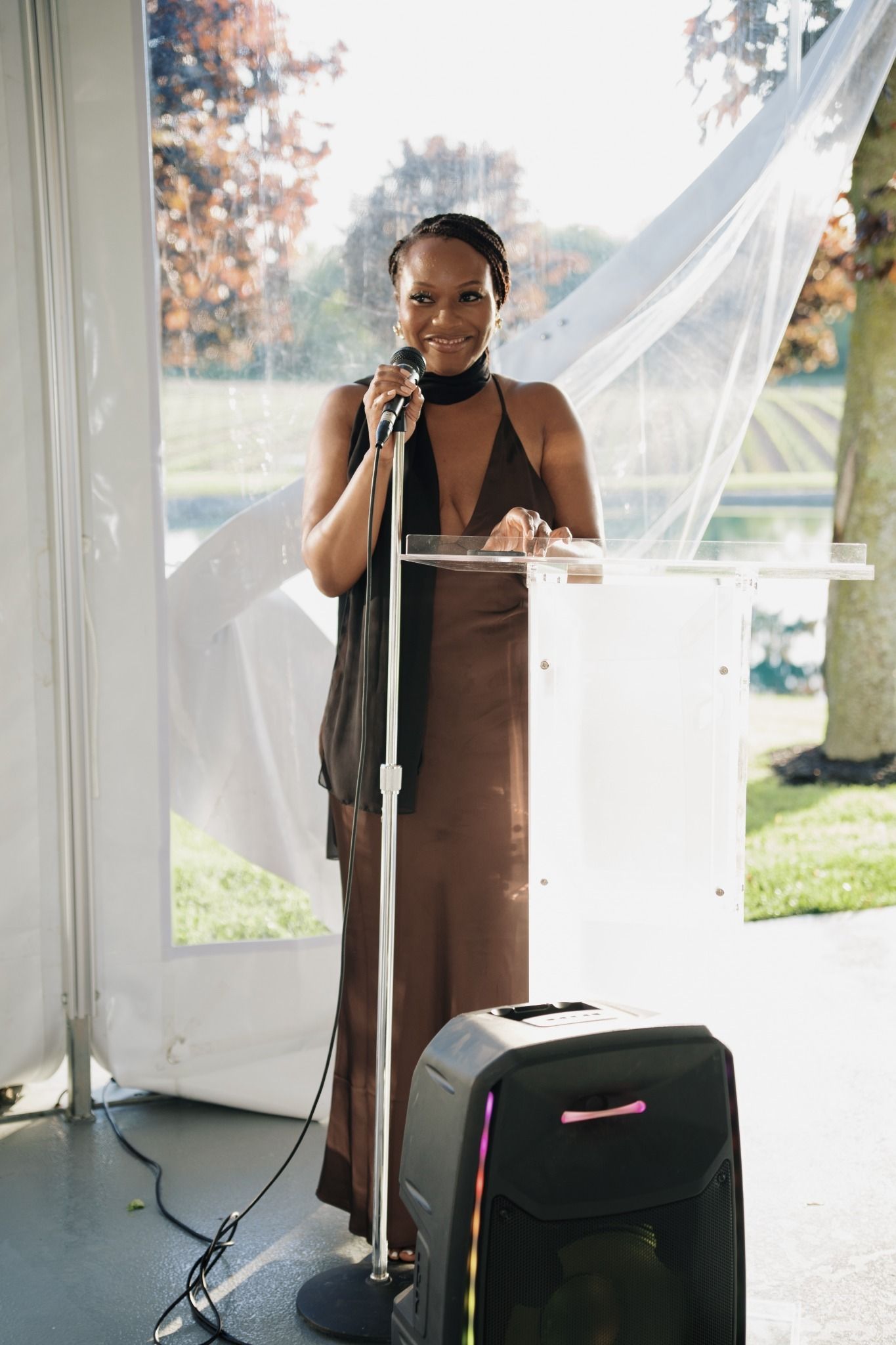 A woman in a brown halter dress speaks at a clear podium with a microphone at an outdoor event.