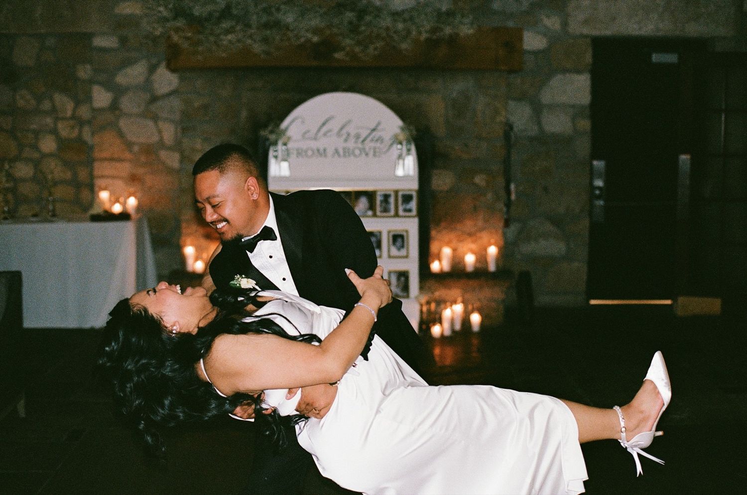 Groom dips bride in elegant wedding reception dance, candlelit stone venue backdrop, bride in white gown with heels.