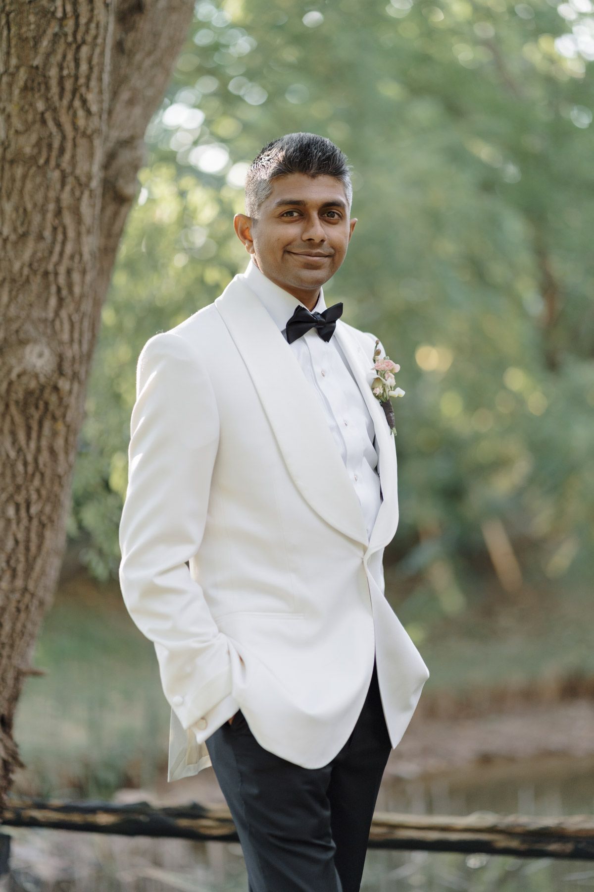 Groom in a white tuxedo jacket and black bow tie posing among green trees at Kurtz Orchards in Niagara-on-the-Lake.