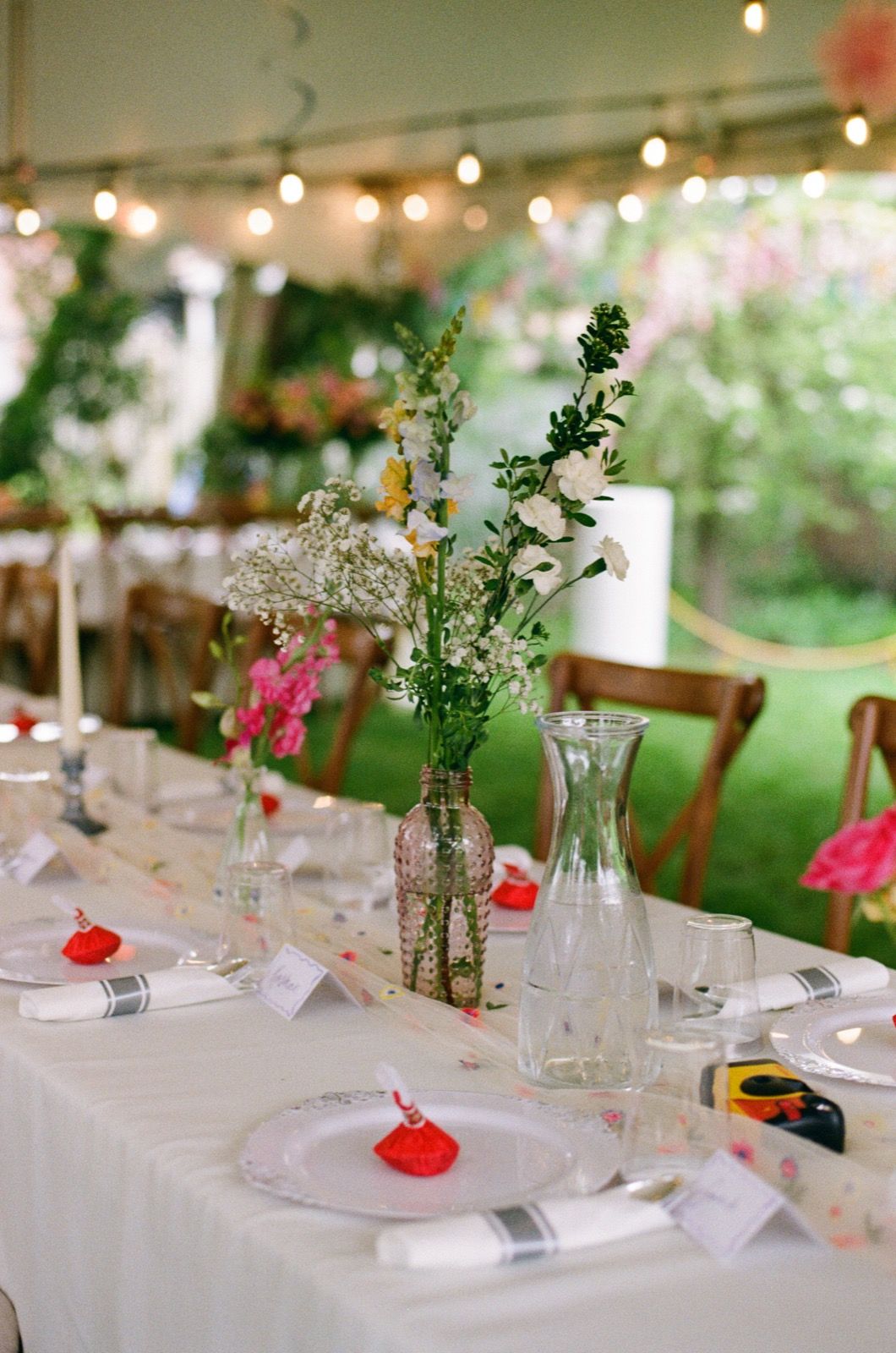 Elegant outdoor wedding reception table with floral centerpieces, string lights, and white linens under a tent.