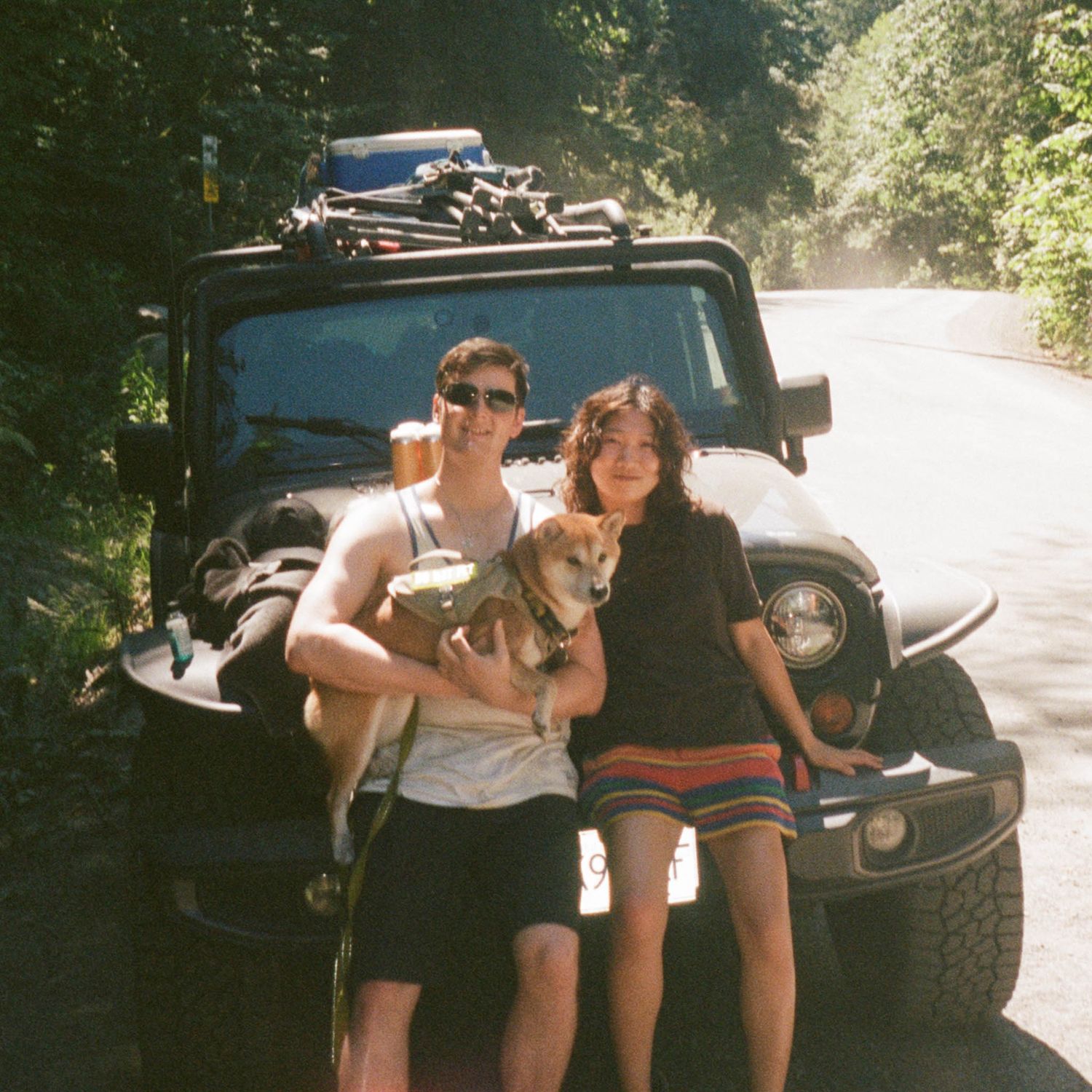 ILO Studio founders Bradley and Clarise with their Shiba Inu Groot posing beside a black Jeep Wrangler on a tree-lined scenic road