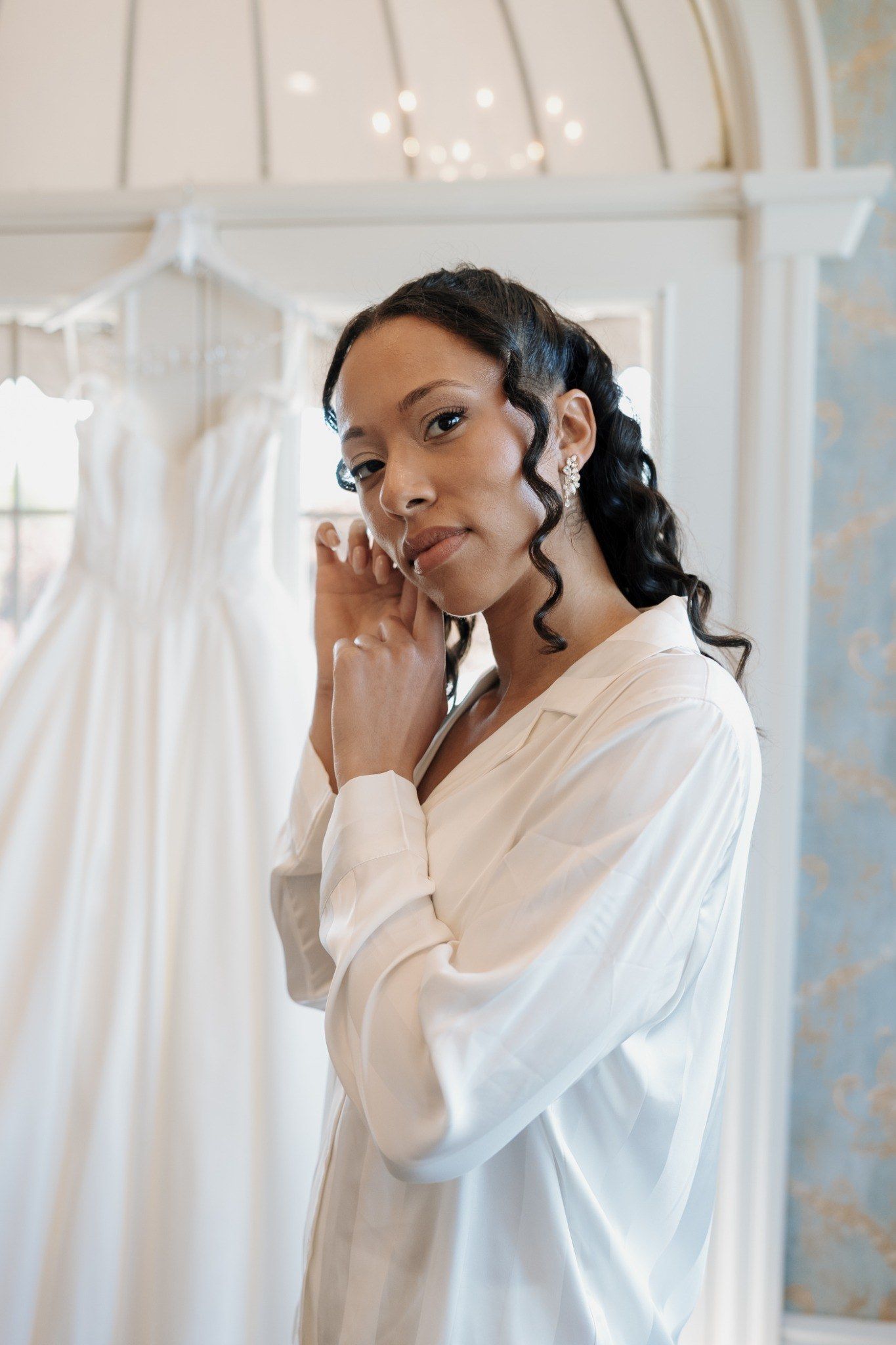 Bride in white silk robe touching her face near wedding dress in elegant bridal suite.