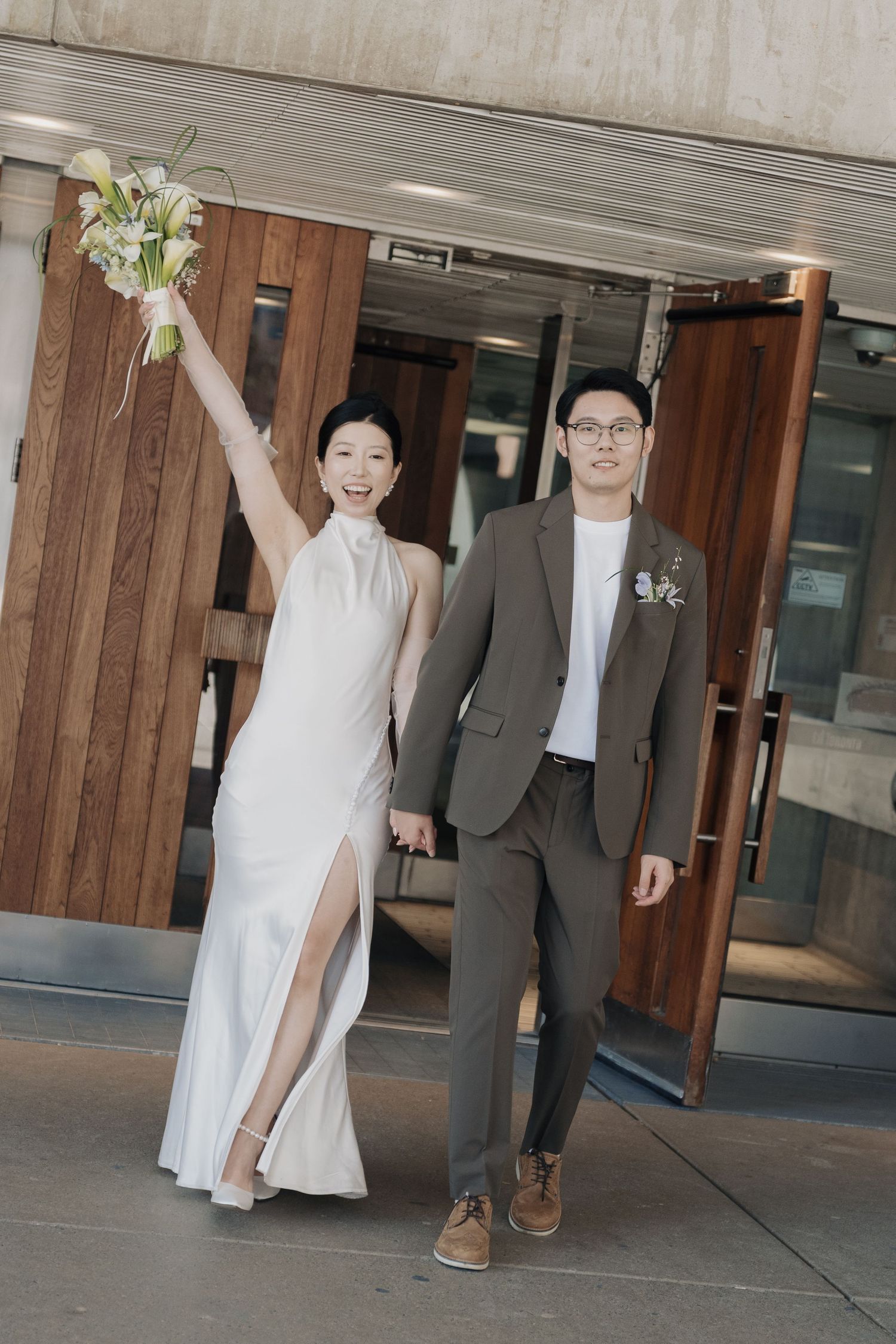 Joyful newlyweds walking out of Toronto City Hall holding hands as bride raises bouquet after their civil wedding ceremony.