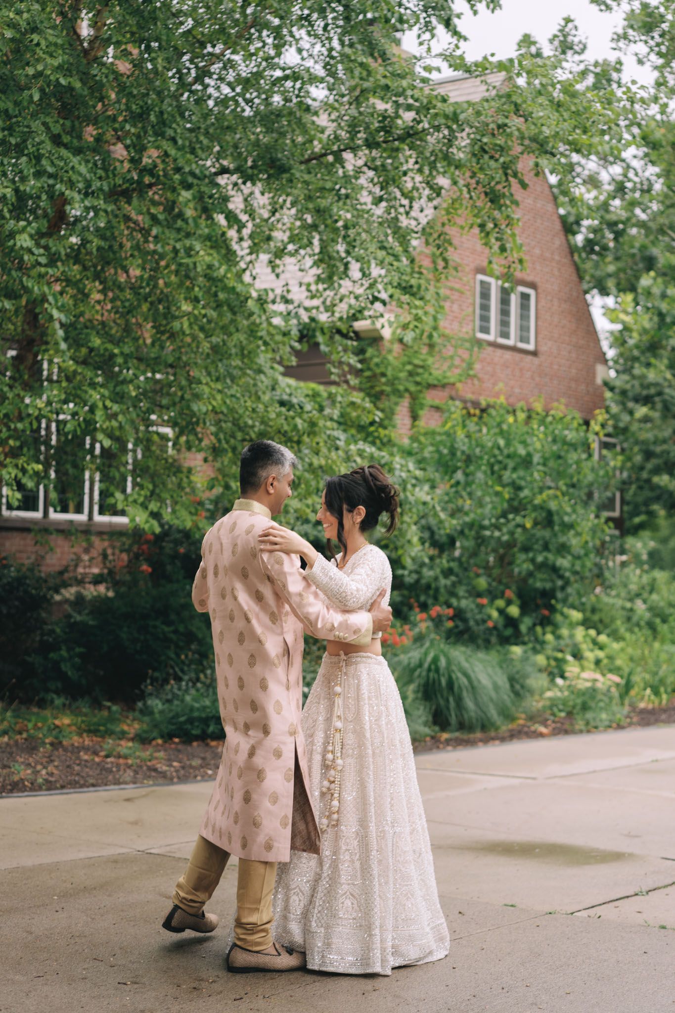 Couple in traditional South Asian wedding attire dancing outdoors near lush greenery and a brick building. McMasters University, Hamilton ON