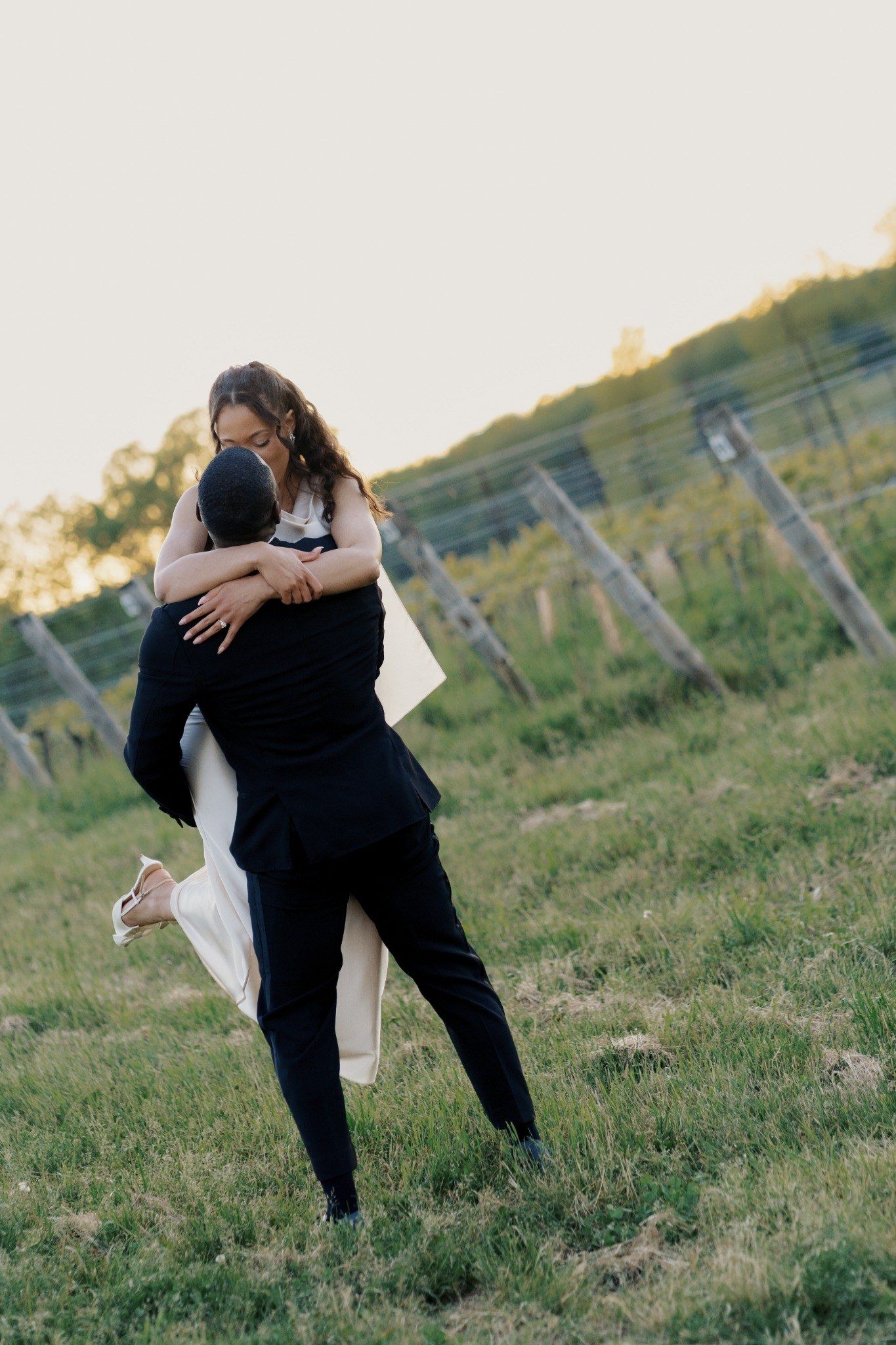 Groom lifting bride in a romantic embrace in a vineyard field at golden hour during their wedding.
