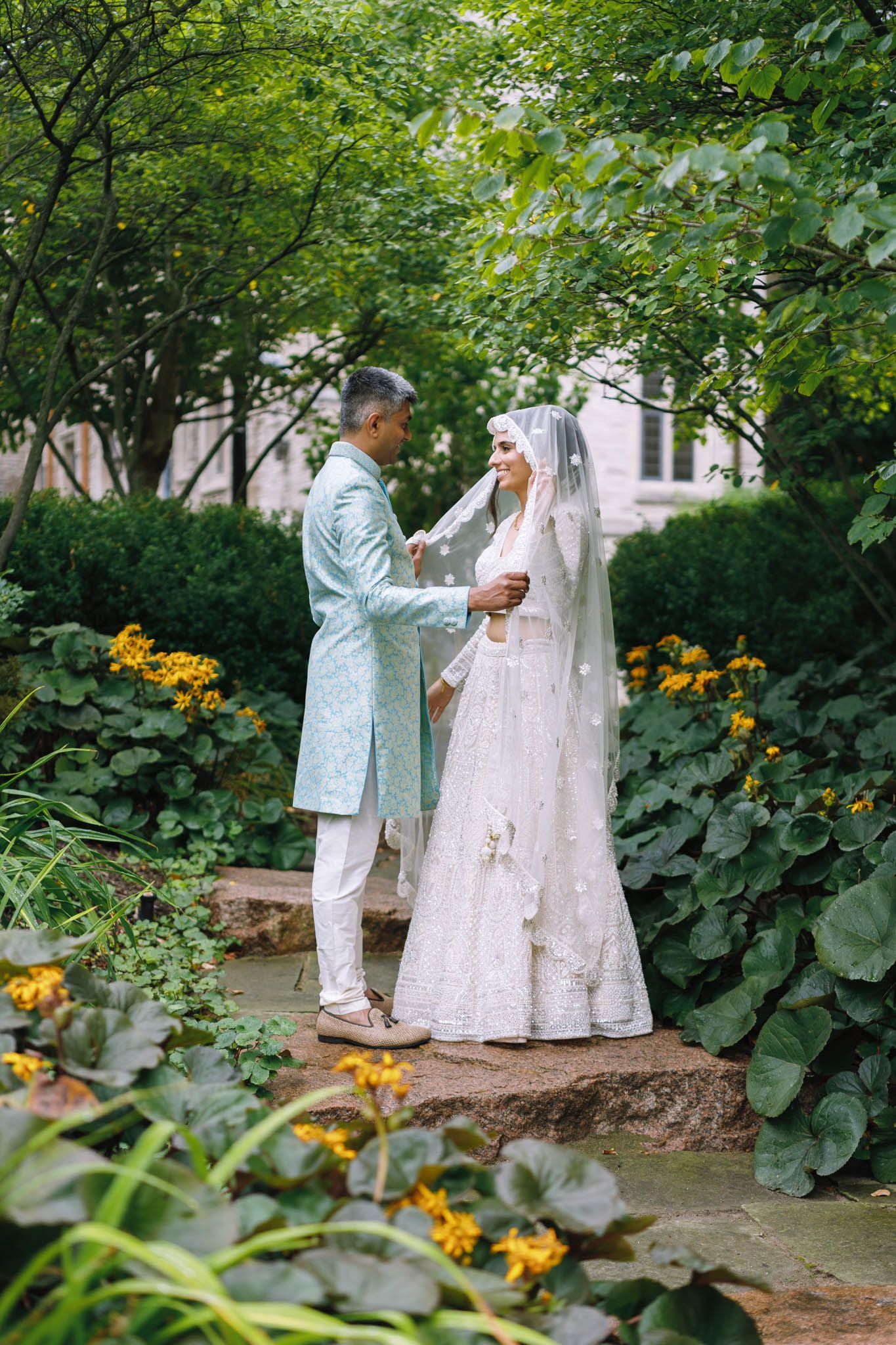 Couple in traditional South Asian wedding attire standing in a lush garden with yellow flowers. McMasters University, Hamilton. Ontario
