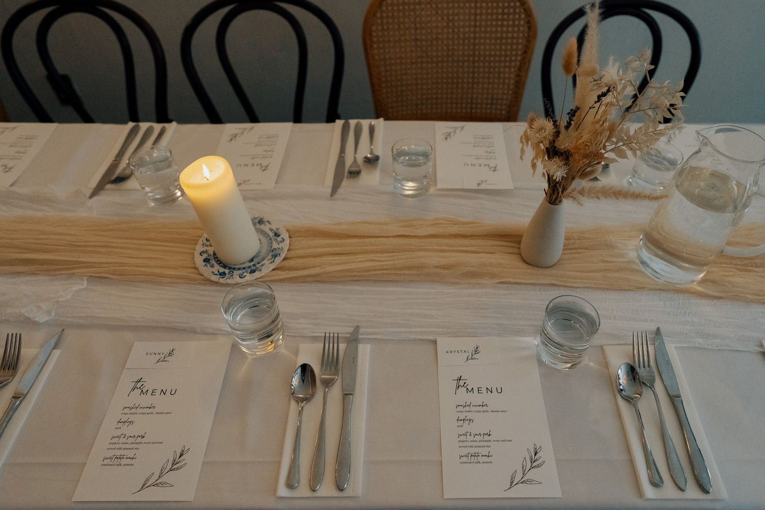 Elegant wedding reception table setting with white linen, candles, dried florals, and printed menus.