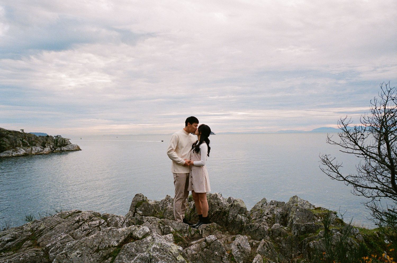 Couple kissing on rocky coastal cliffs, calm blue ocean and cloudy sky at Whytecliff Park in West Vancouver on 35mm film engagement