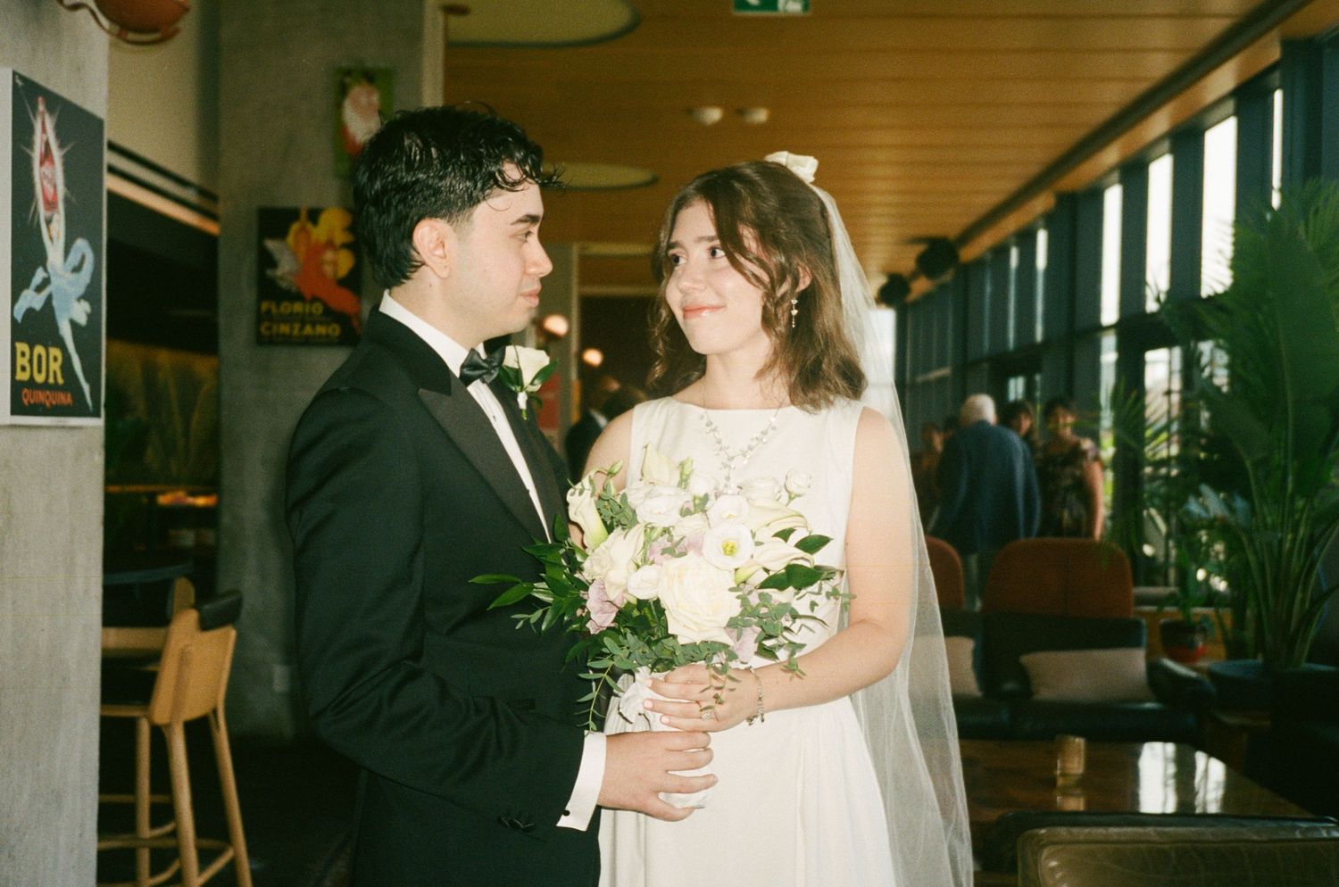 Bride in white gown holding bouquet stands with groom in black tuxedo at indoor wedding venue.