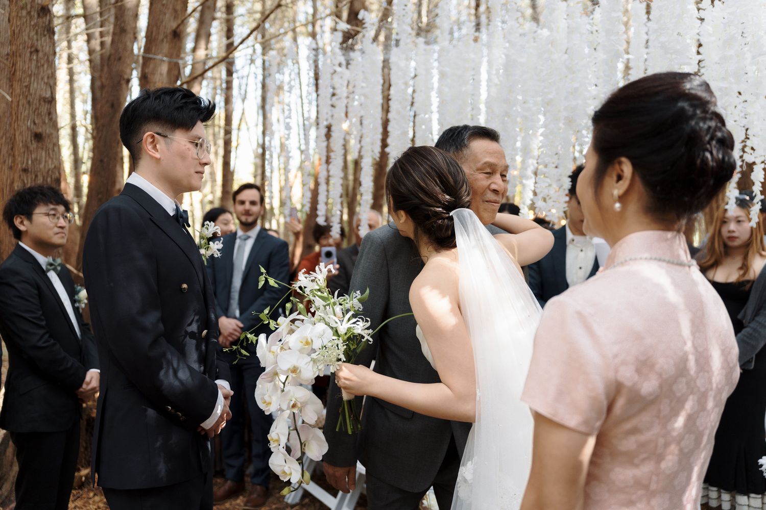 Asian couple exchanging vows with white orchids and family nearby at Razi Estate in Schomberg for forest wedding photography.