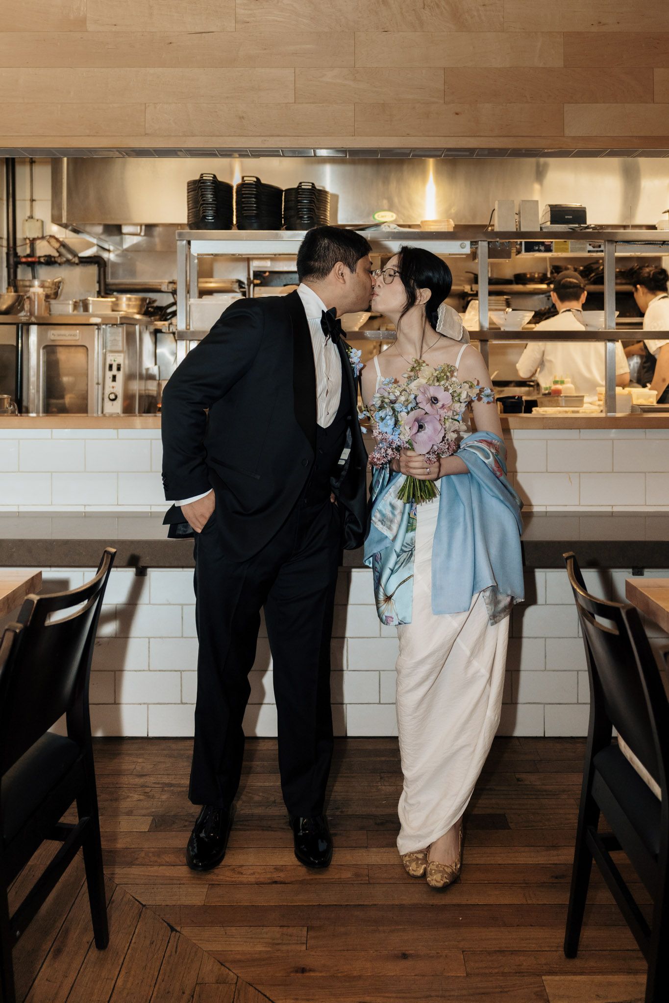 Newlyweds share a kiss in a restaurant kitchen, groom in tuxedo, bride in white gown with blue sash holding floral bouquet.