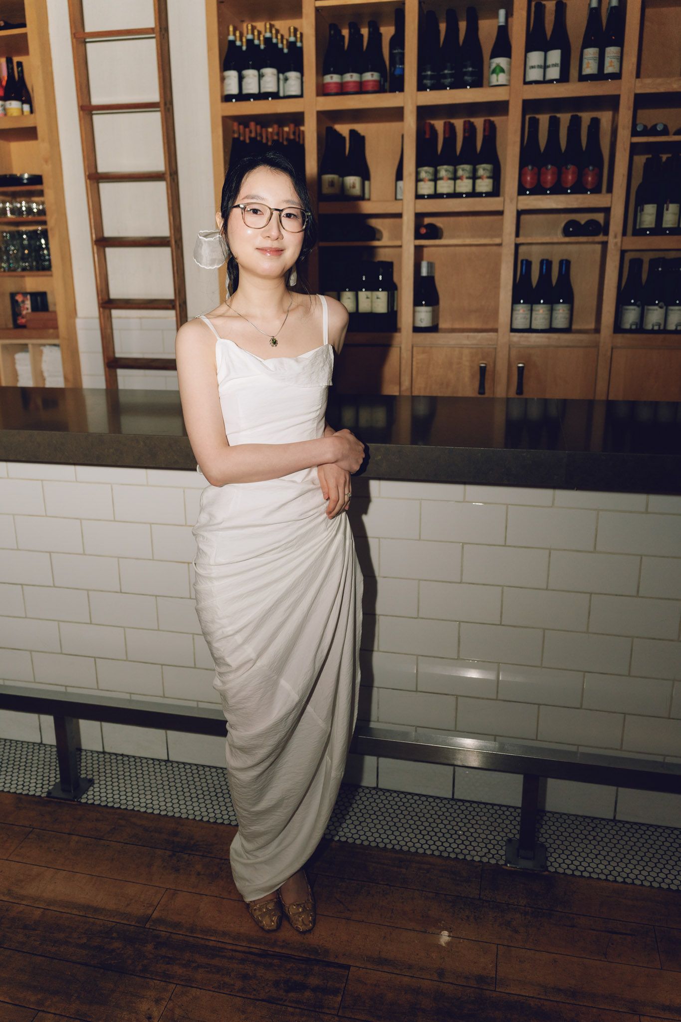 Woman in white dress and glasses posing elegantly in a wine bar with bottle-lined shelves.