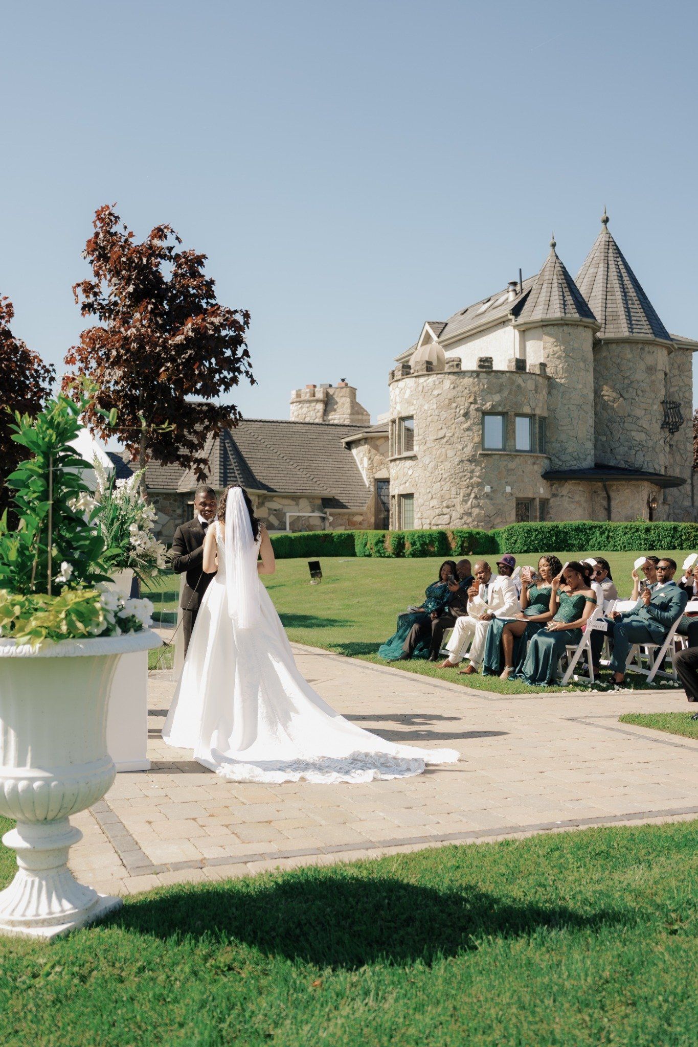 Bride in white gown with veil stands at outdoor wedding ceremony before stone castle venue on sunny day.