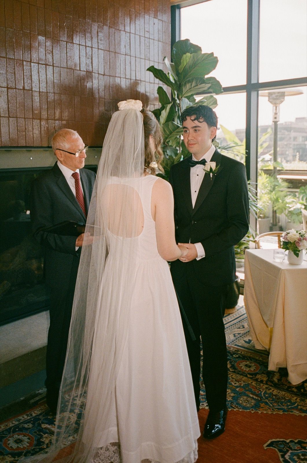Bride and groom exchange vows at indoor wedding ceremony with officiant, bride wearing white gown and veil, groom in black tuxedo.