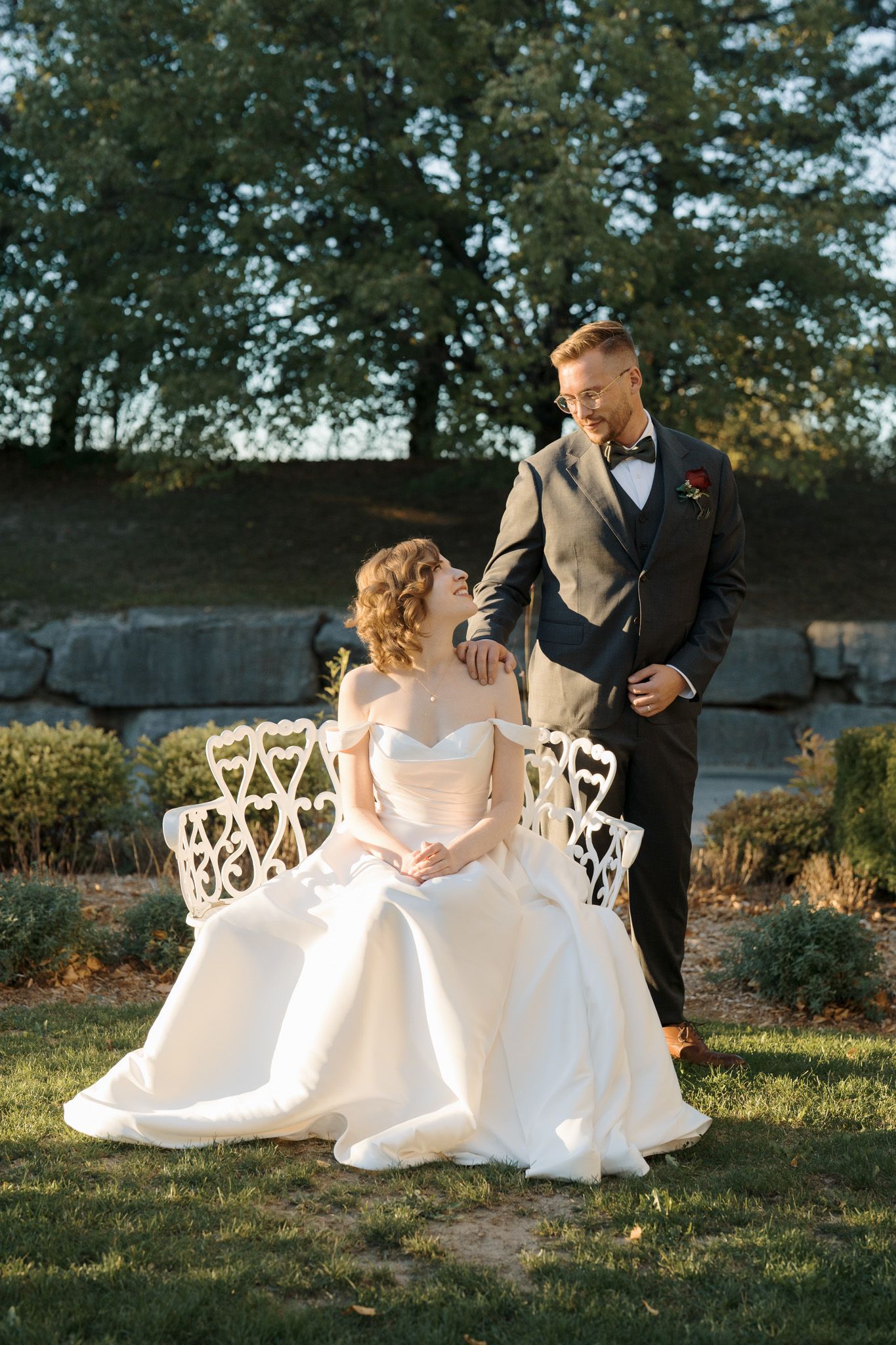 Bride seated on an ornate bench as groom stands beside her in a sunlit garden at The Manor during their Jewish wedding portraits.