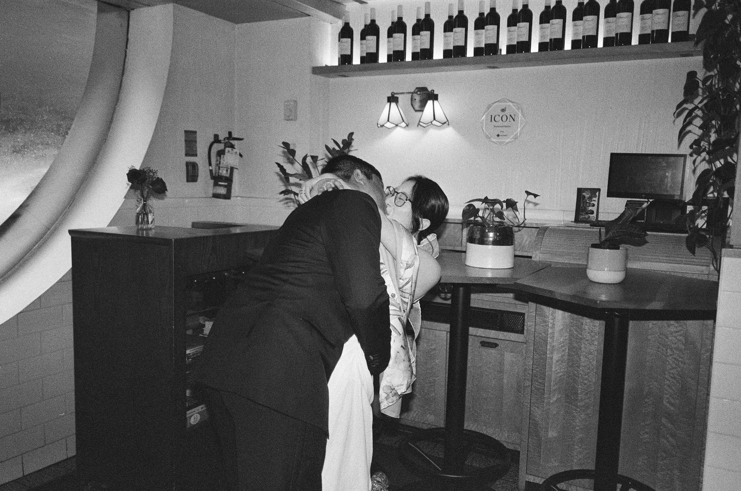 Black and white photo of a couple sharing a romantic moment at a bar counter with wine bottles displayed on shelves.