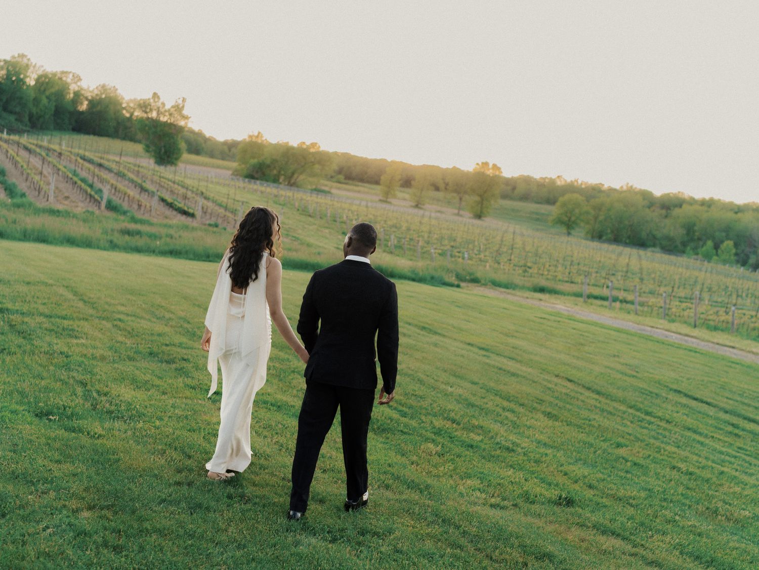 Bride and groom walking hand in hand through a vineyard at golden hour sunset.