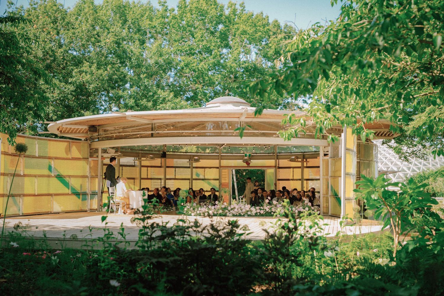 Outdoor wedding ceremony under a pavilion surrounded by lush green trees in summer.