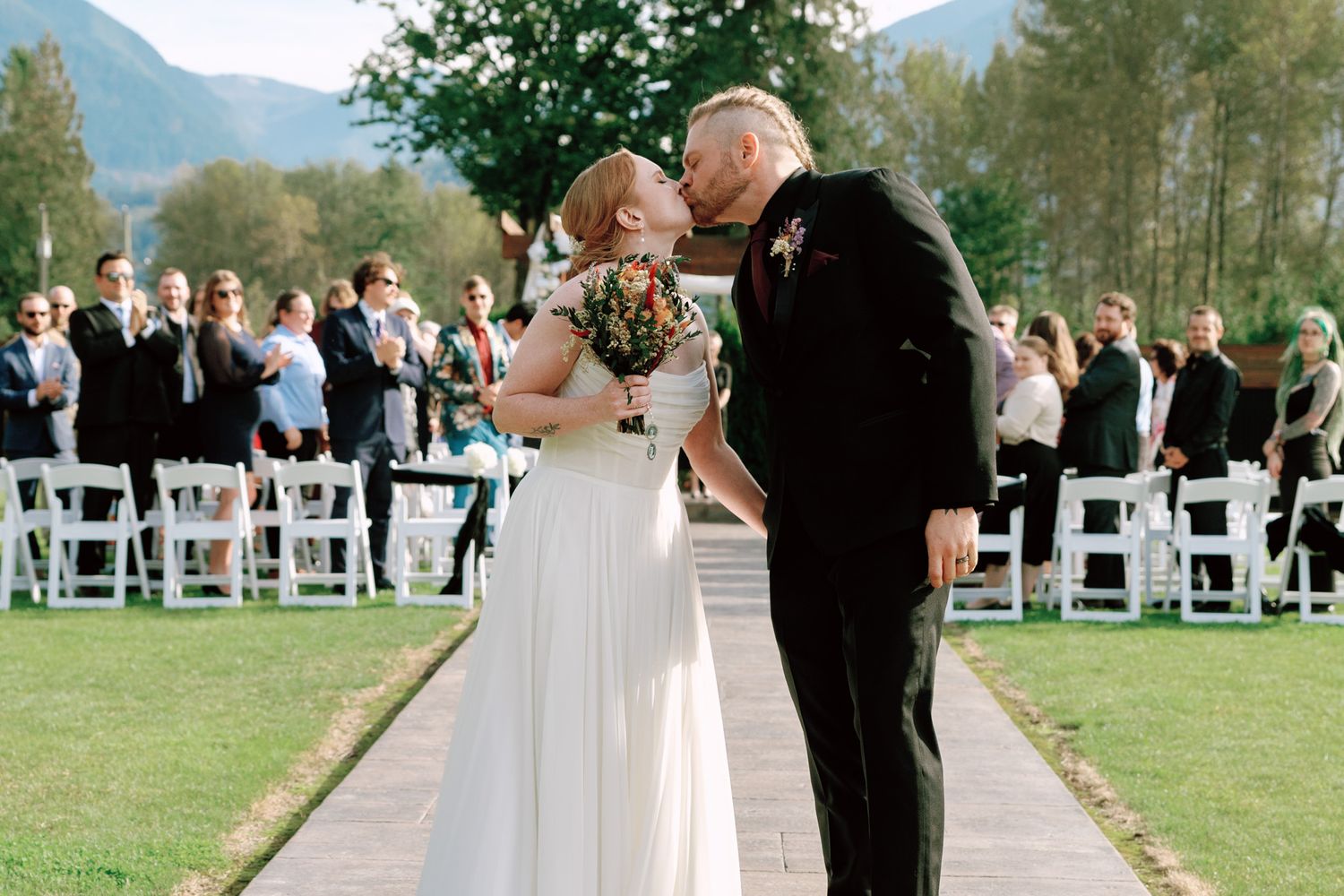 Bride and groom share a first kiss at outdoor wedding ceremony with white chairs and mountain backdrop.