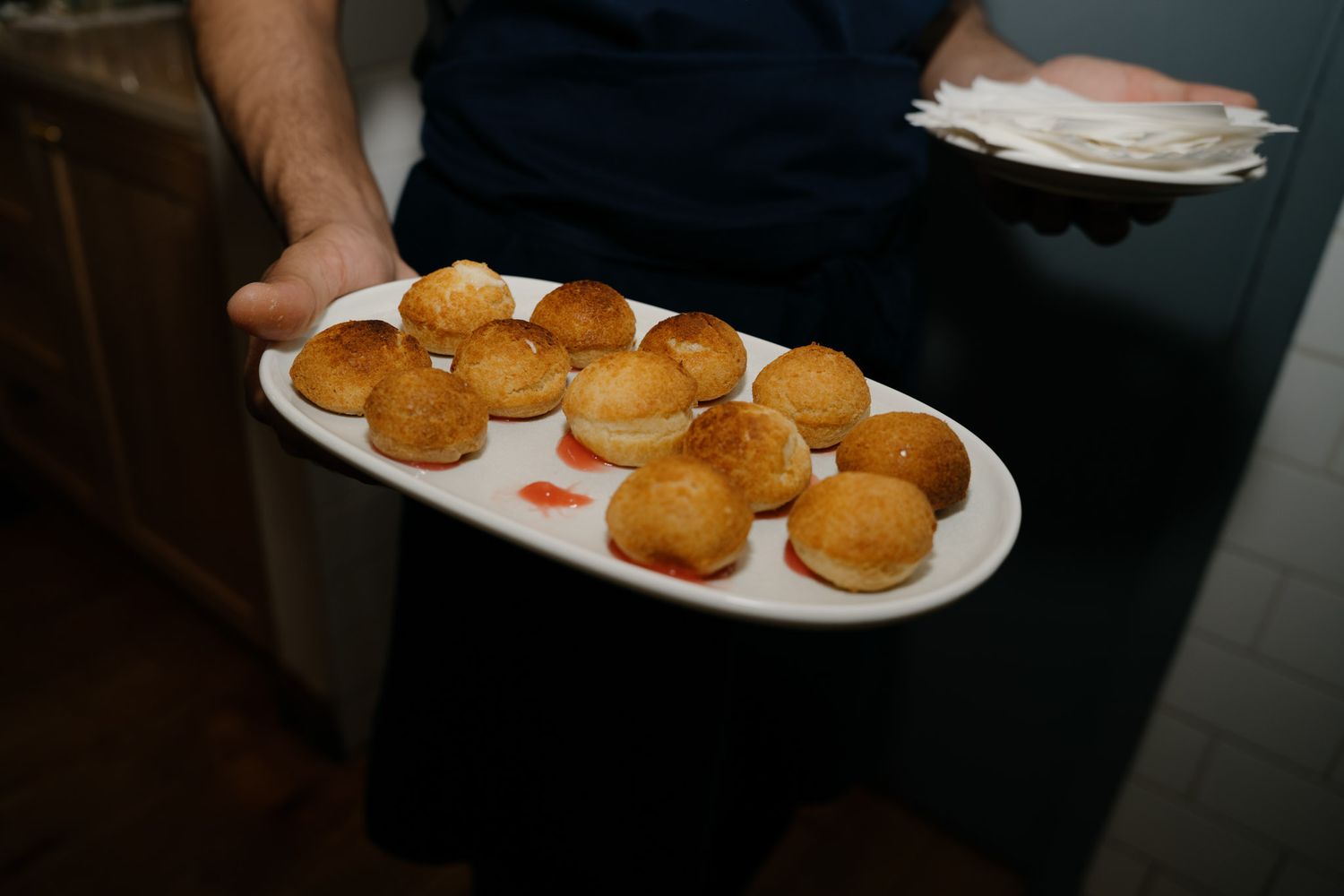 Waiter serving a white oval platter of golden baked bread rolls with red sauce in a restaurant setting.