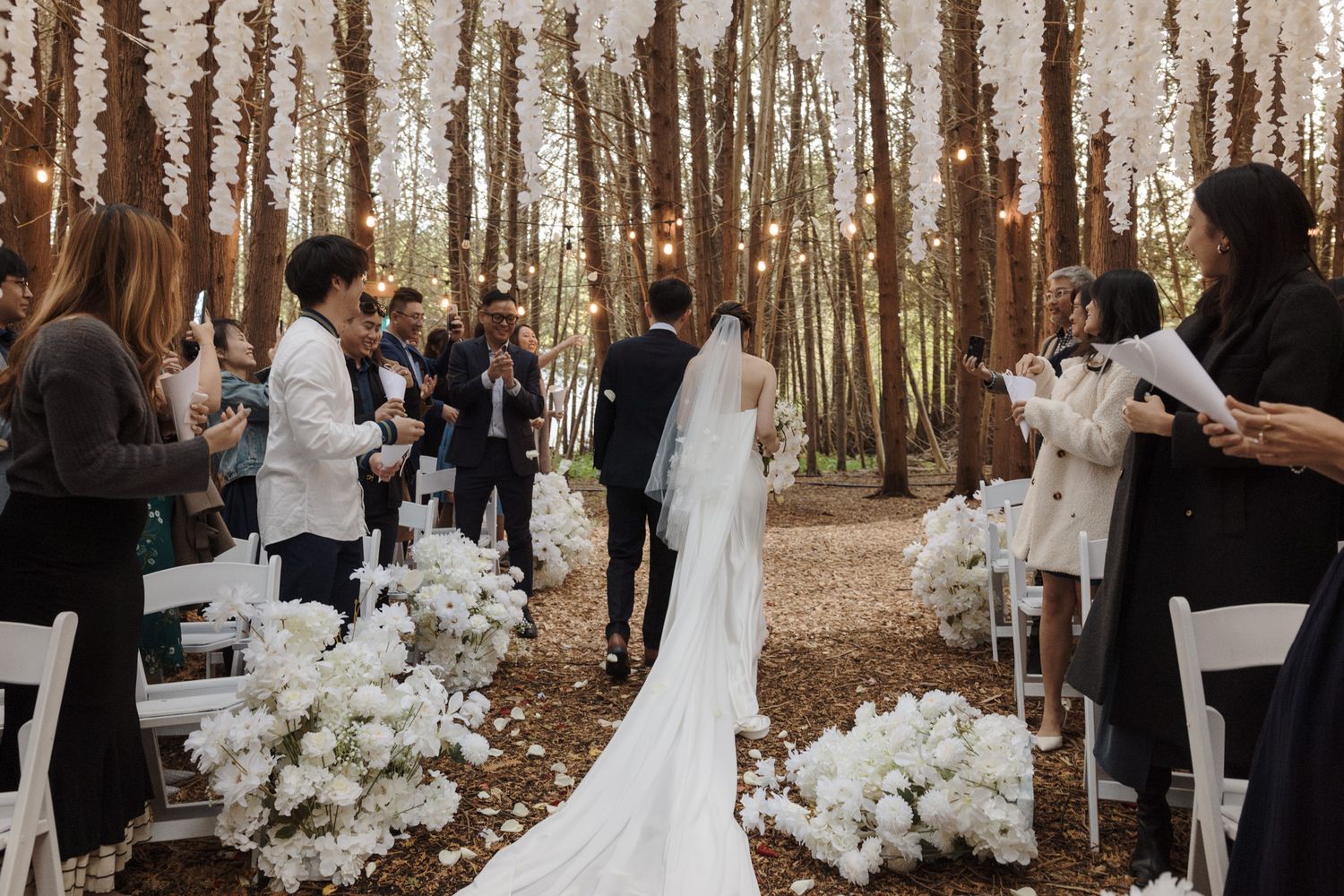 Bride walking down a forest aisle with white florals and hanging lights at Razi Estate in Schomberg for woodland wedding photography.