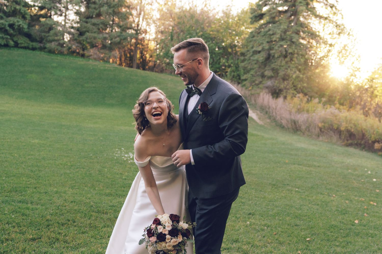 Bride and groom laughing on a green lawn at golden hour, bride holding a burgundy bouquet, during their Jewish wedding at the Manor, Ontario