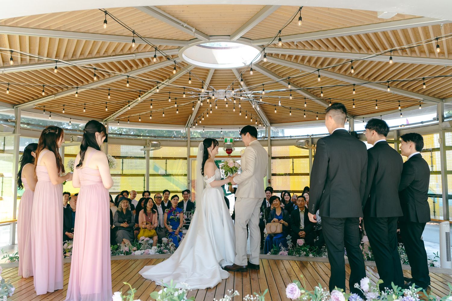 Asian couple exchanges vows under a wooden domed pavilion with string lights and pink bridesmaids.