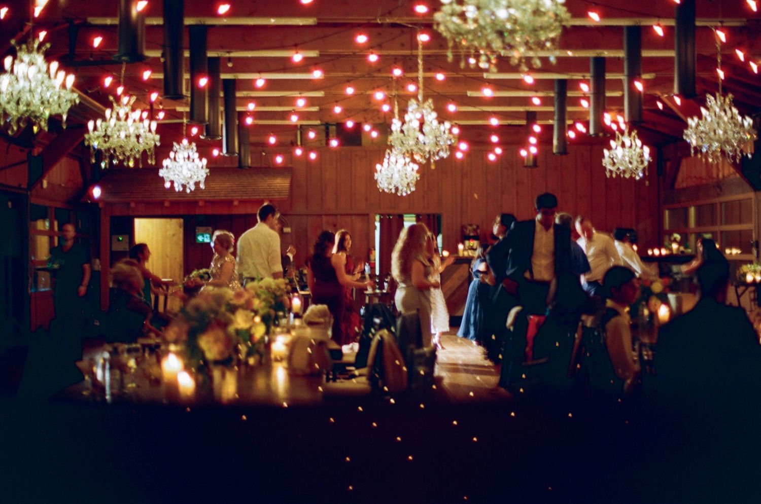 Elegant wedding reception venue with crystal chandeliers, string lights, and guests mingling in a warmly lit rustic ballroom.