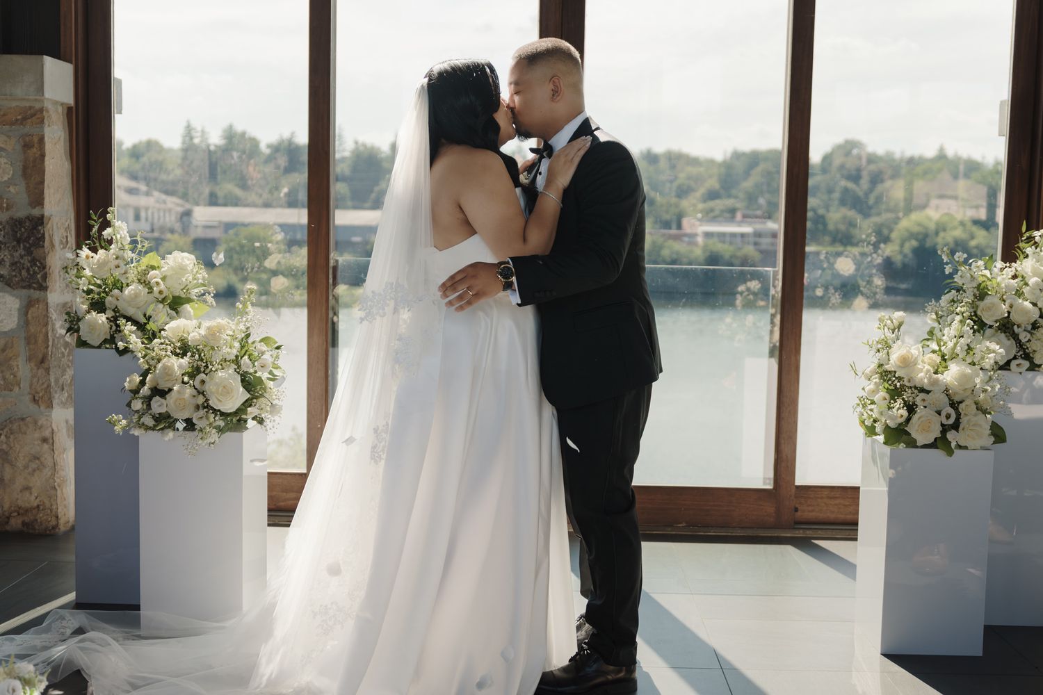 Bride and groom sharing their first kiss with white florals and waterfront views at Cambridge Mill for elegant wedding photography.