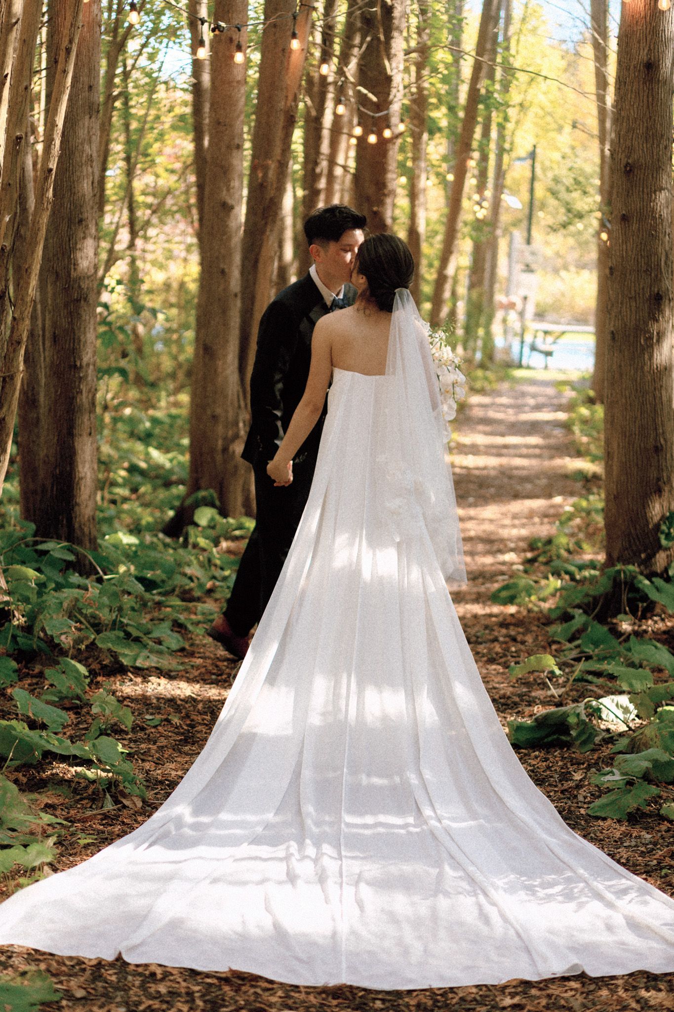 Bride and groom kissing on a forest path with cathedral veil trailing at Razi Estate in Schomberg for cinematic wedding photography.