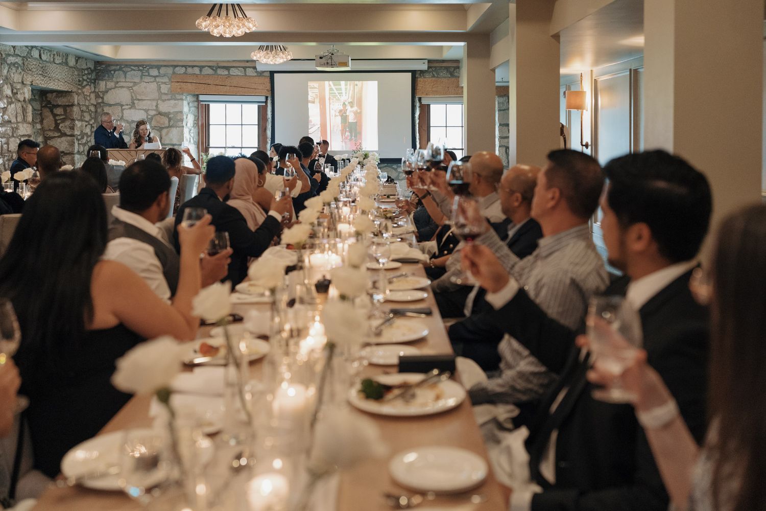 Wedding guests raise their glasses in a toast at an elegant long reception dinner table.
