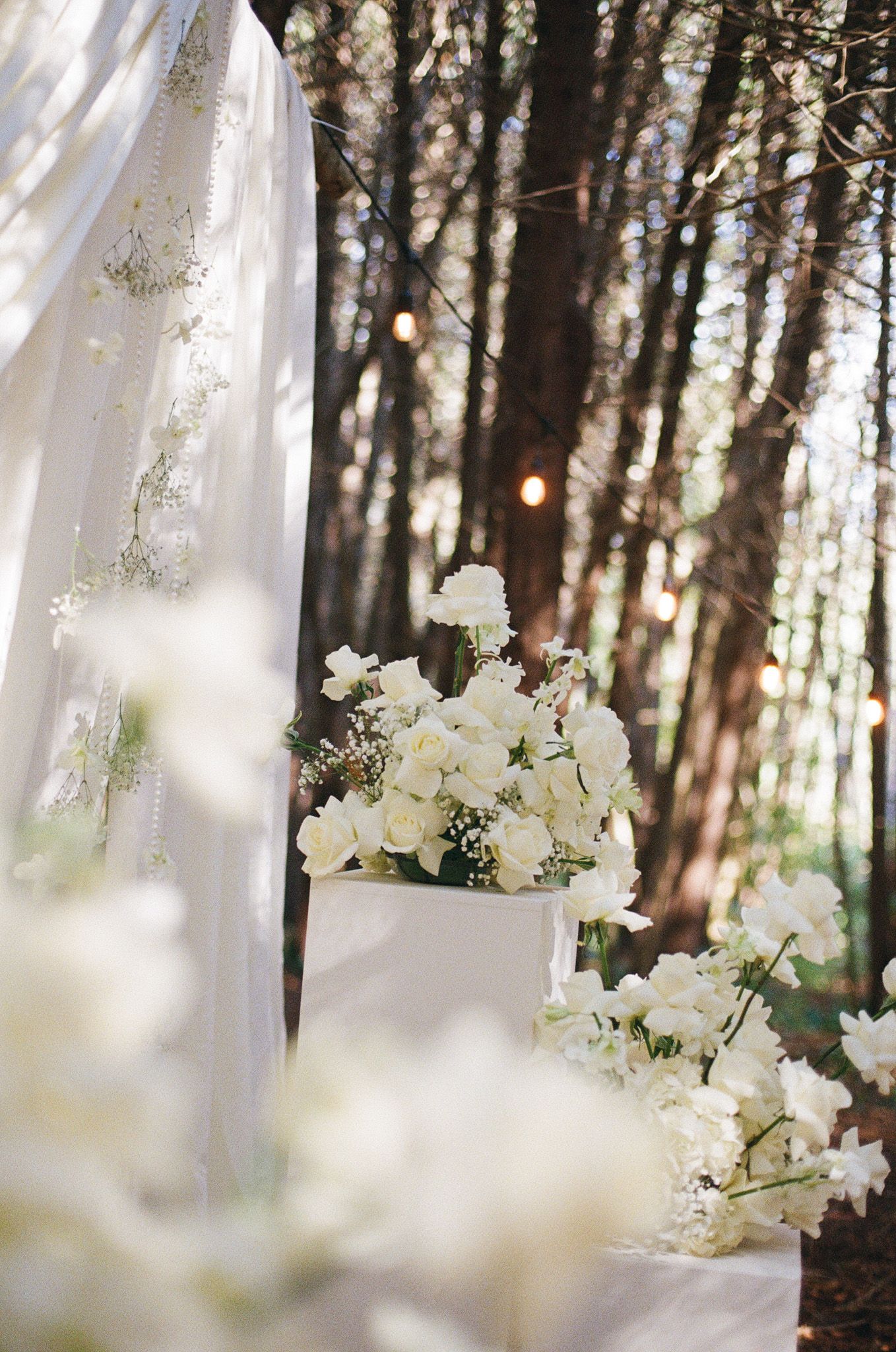 White roses and baby’s breath on a ceremony pedestal with sheer draping at Razi Estate in Schomberg for forest wedding photography.