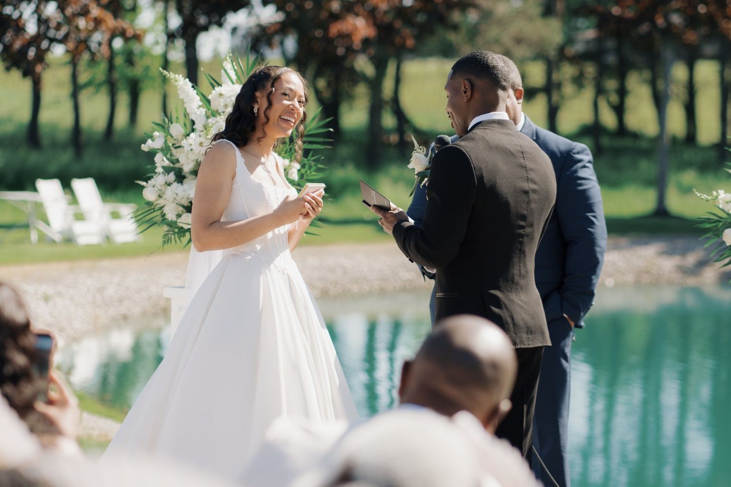 Bride in white gown laughing with groom in navy suit during outdoor wedding ceremony by a serene green pond.