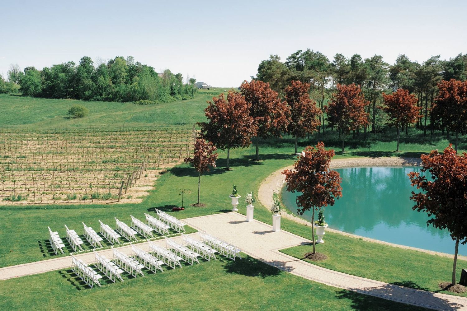 Aerial view of an elegant outdoor wedding ceremony setup with white chairs beside a serene pond at a vineyard.