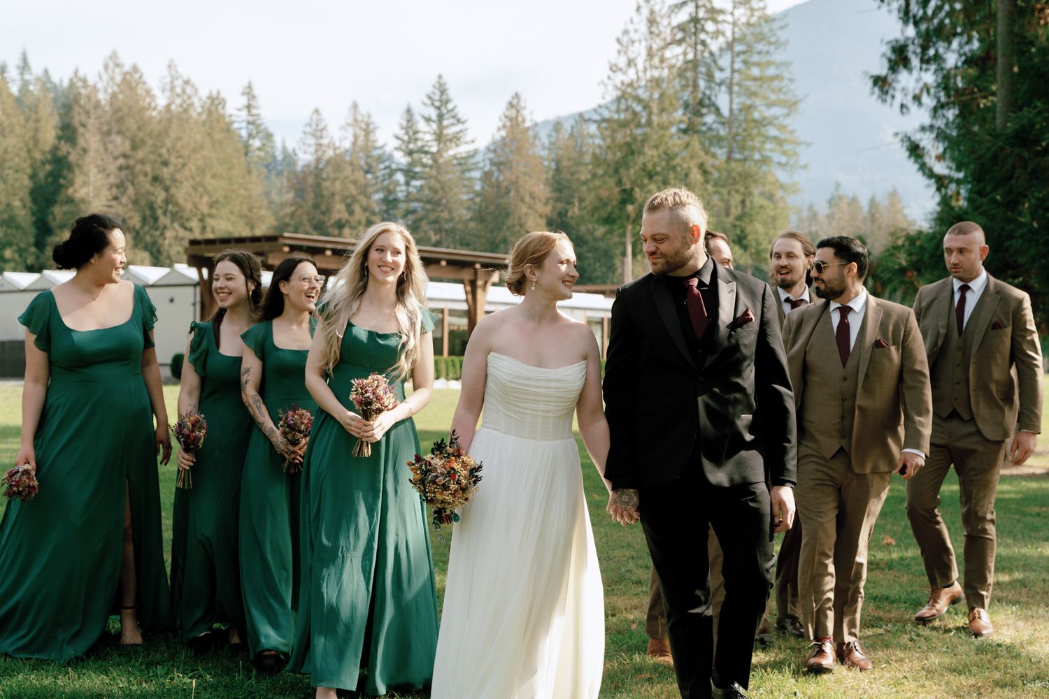Wedding party walks together outdoors with bridesmaids in emerald green dresses and groomsmen in brown suits.