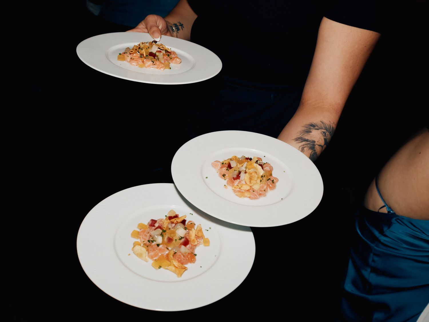Tattooed servers presenting three elegant white plates with gourmet pasta dishes at a fine dining restaurant.