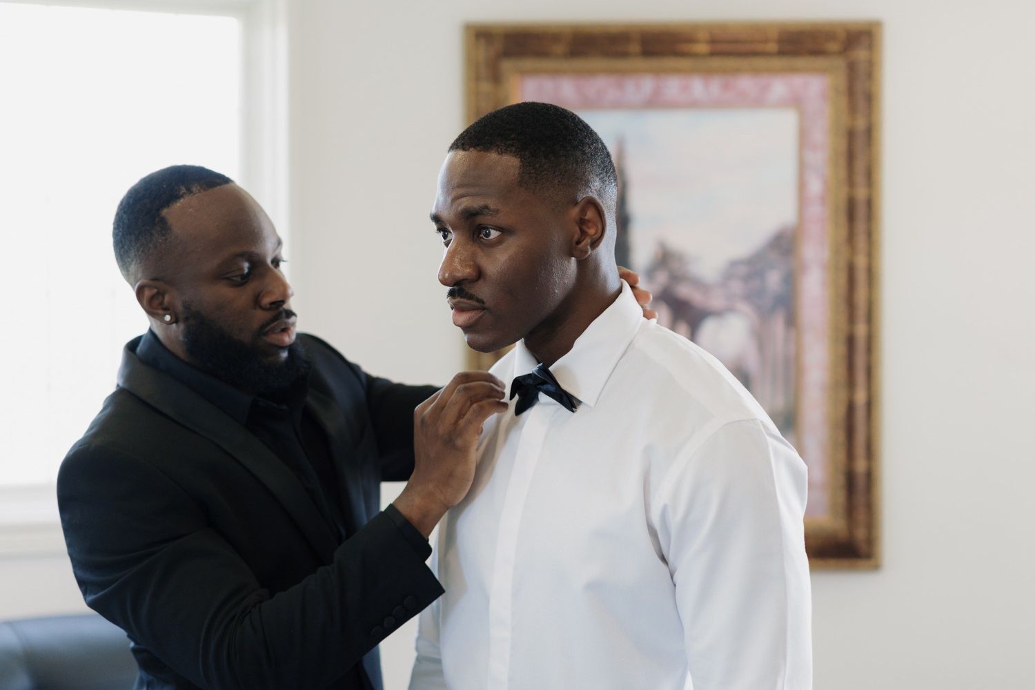 A man in a black suit adjusts a bow tie on another man in a white dress shirt, preparing for a formal event.
