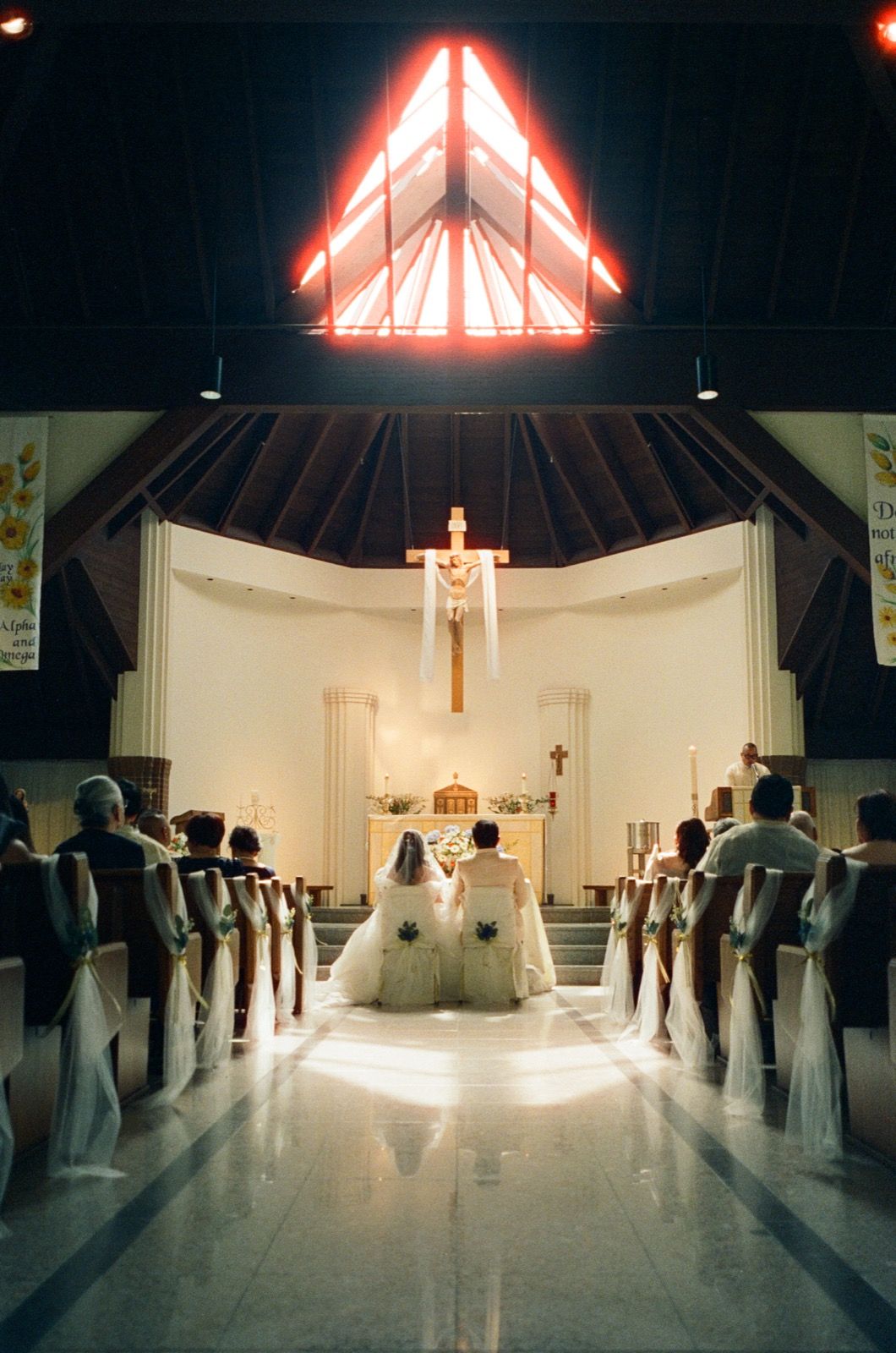 A wedding ceremony in a church with couple at the altar, wooden pews, and a glowing red triangular window above.
