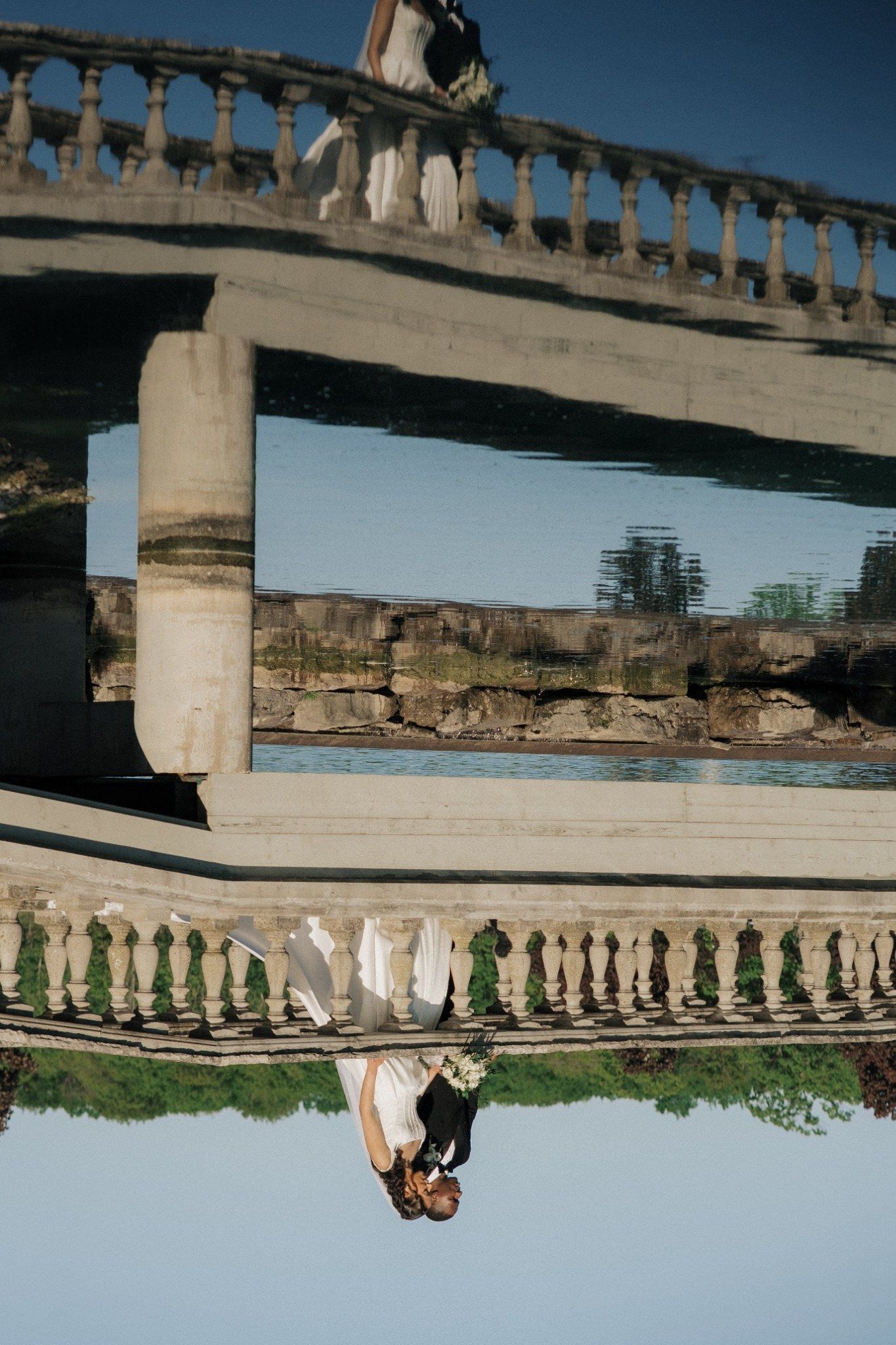 Bride and groom reflected upside down under a stone bridge over a river in a creative wedding photo.