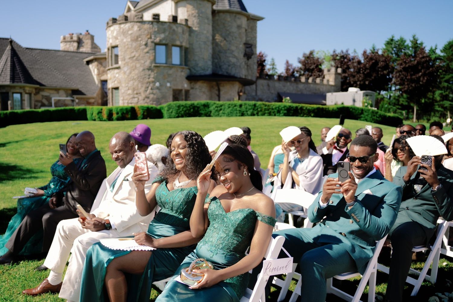 Elegant outdoor wedding ceremony at a castle venue with guests in teal formal attire seated on white chairs.