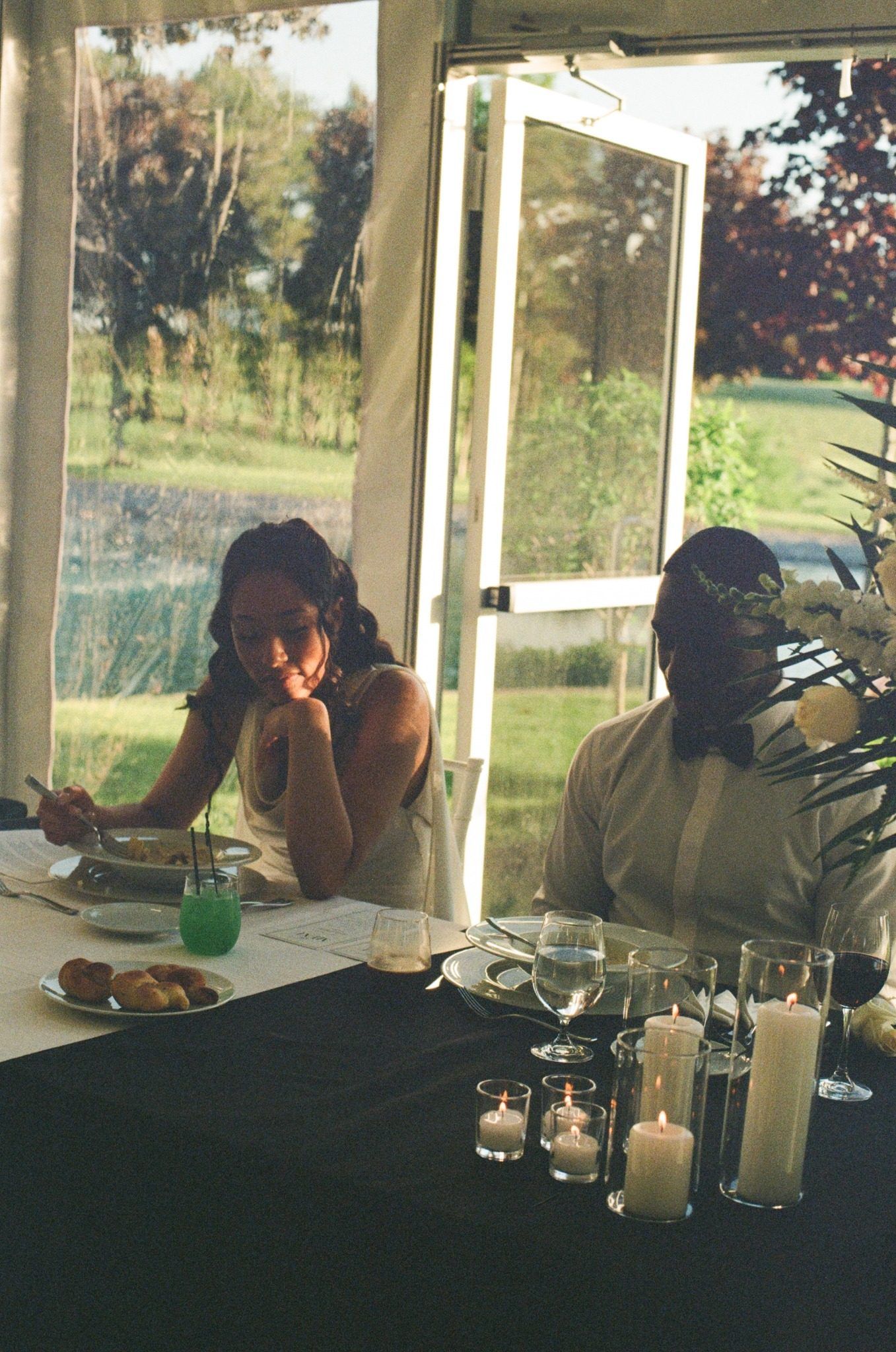Two women sit at a candlelit wedding reception table near large windows overlooking a green outdoor landscape.