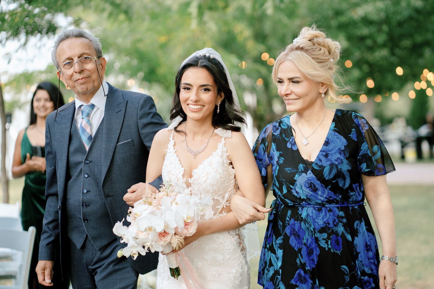 Bride walking with parents beneath string lights at Kurtz Orchards in Niagara-on-the-Lake during an elegant outdoor wedding ceremony.