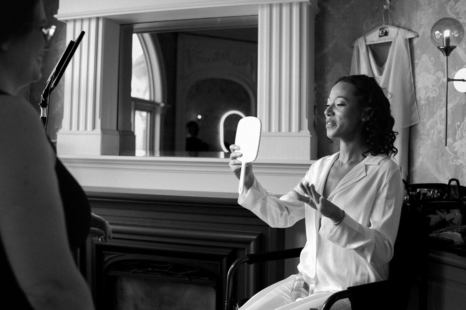 Bride in white robe holds hand mirror while getting ready, captured in elegant black and white photography.