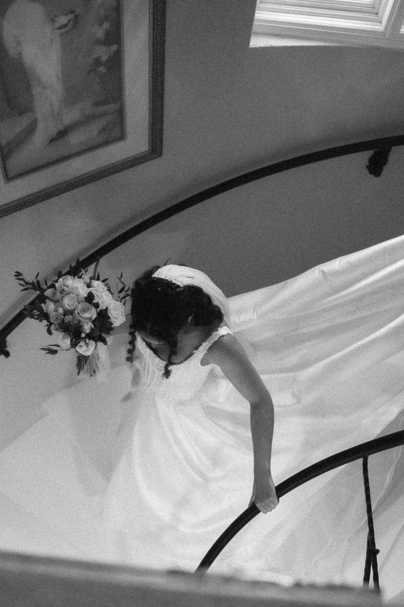 Black and white photo of a bride in a flowing white gown descending a curved staircase while holding a bouquet.