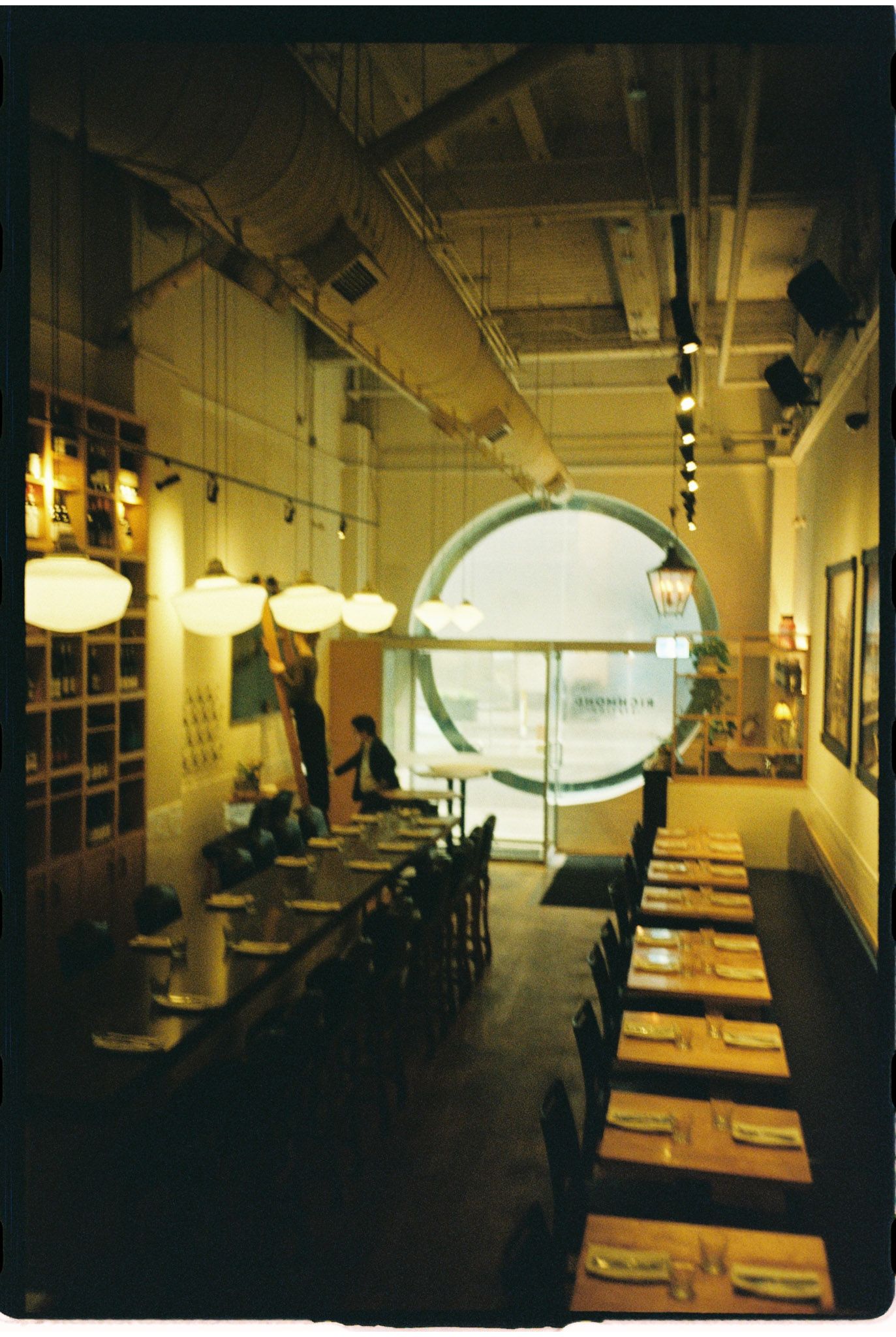Cozy restaurant interior with long dining tables, pendant lights, and a circular window.