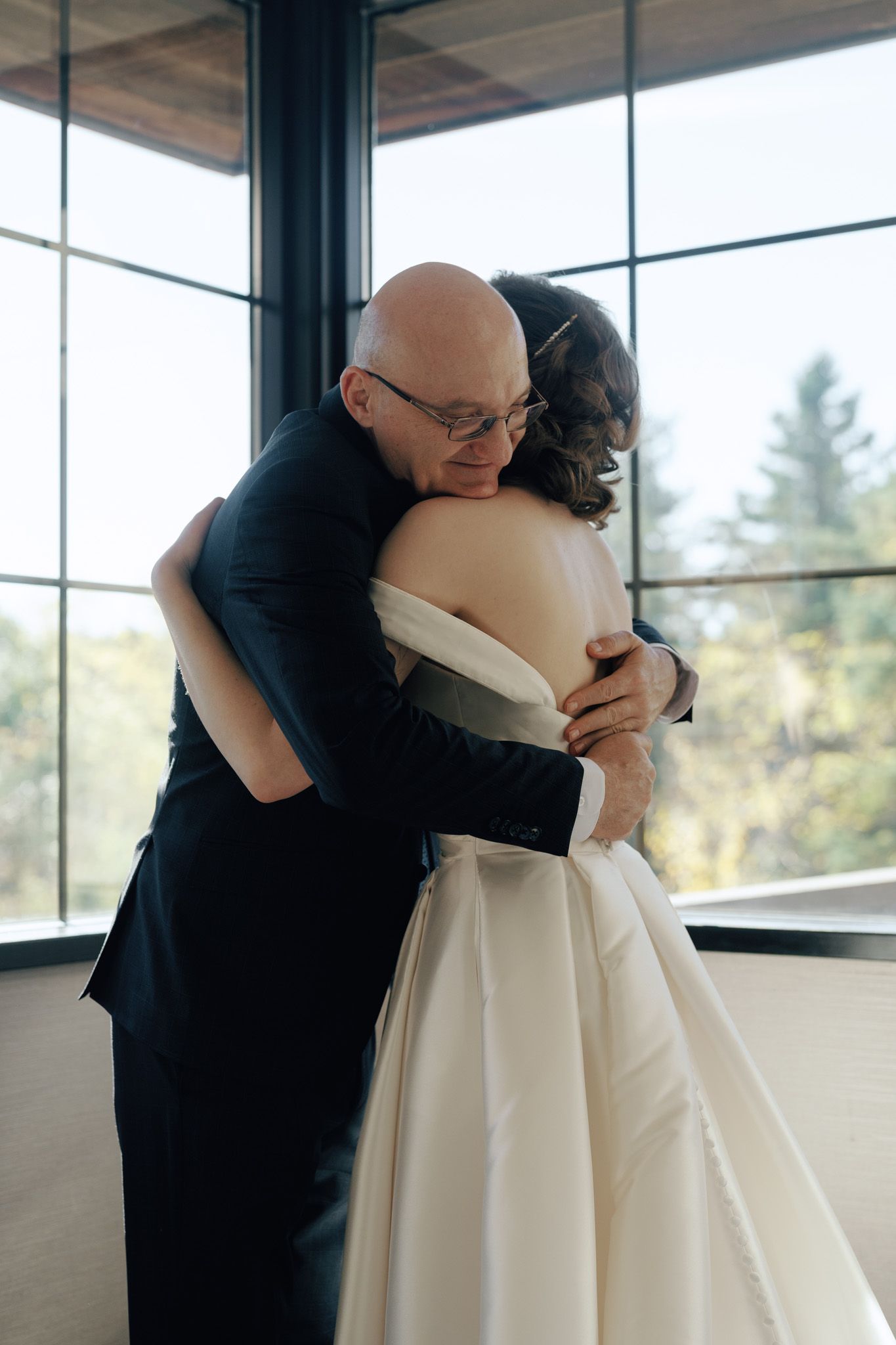 Bride sharing an emotional hug with a man in a dark suit by bright windows at The Manor in King City wedding morning.