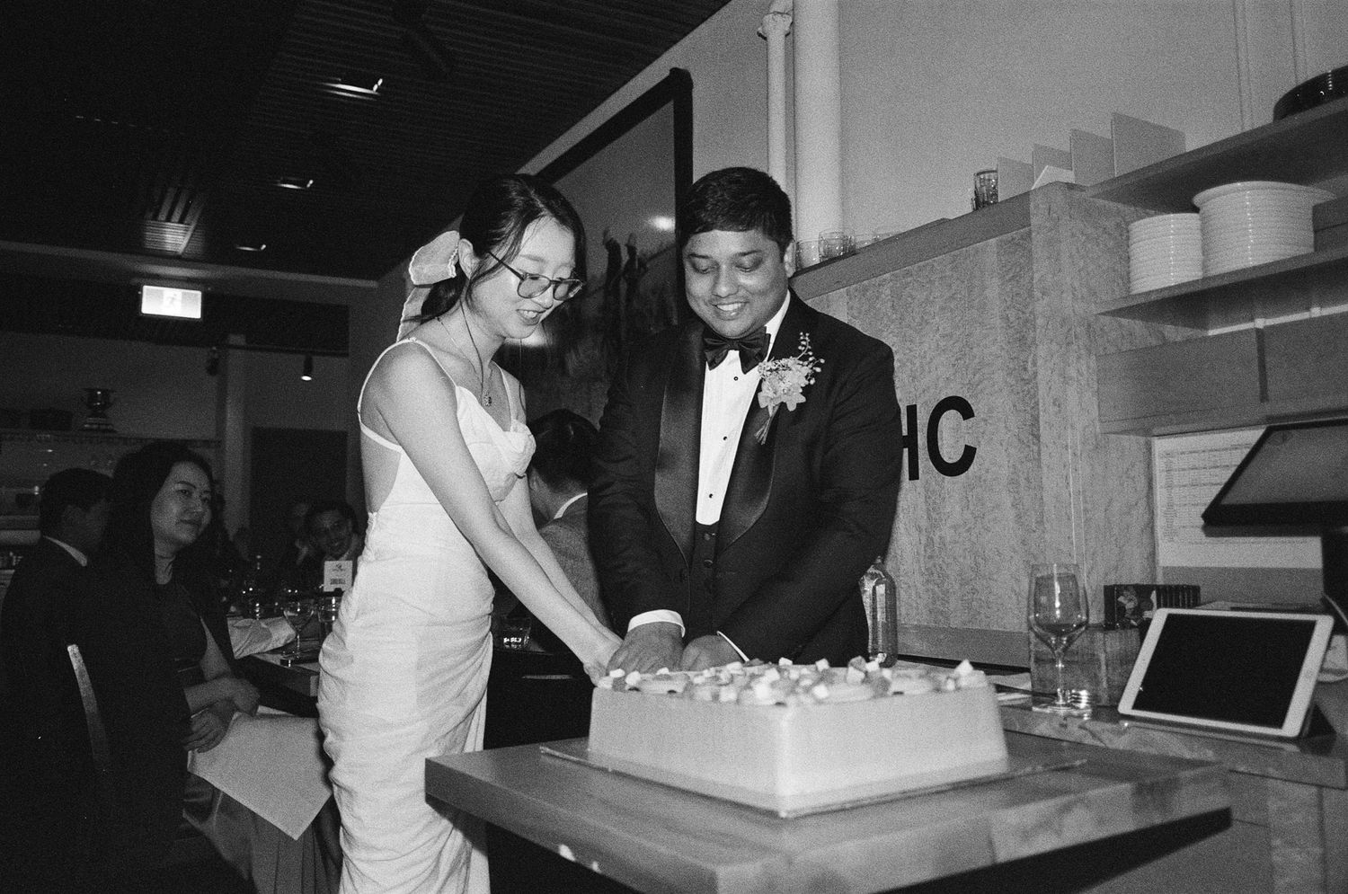 Couple cutting wedding cake together in black and white photo at indoor reception venue.